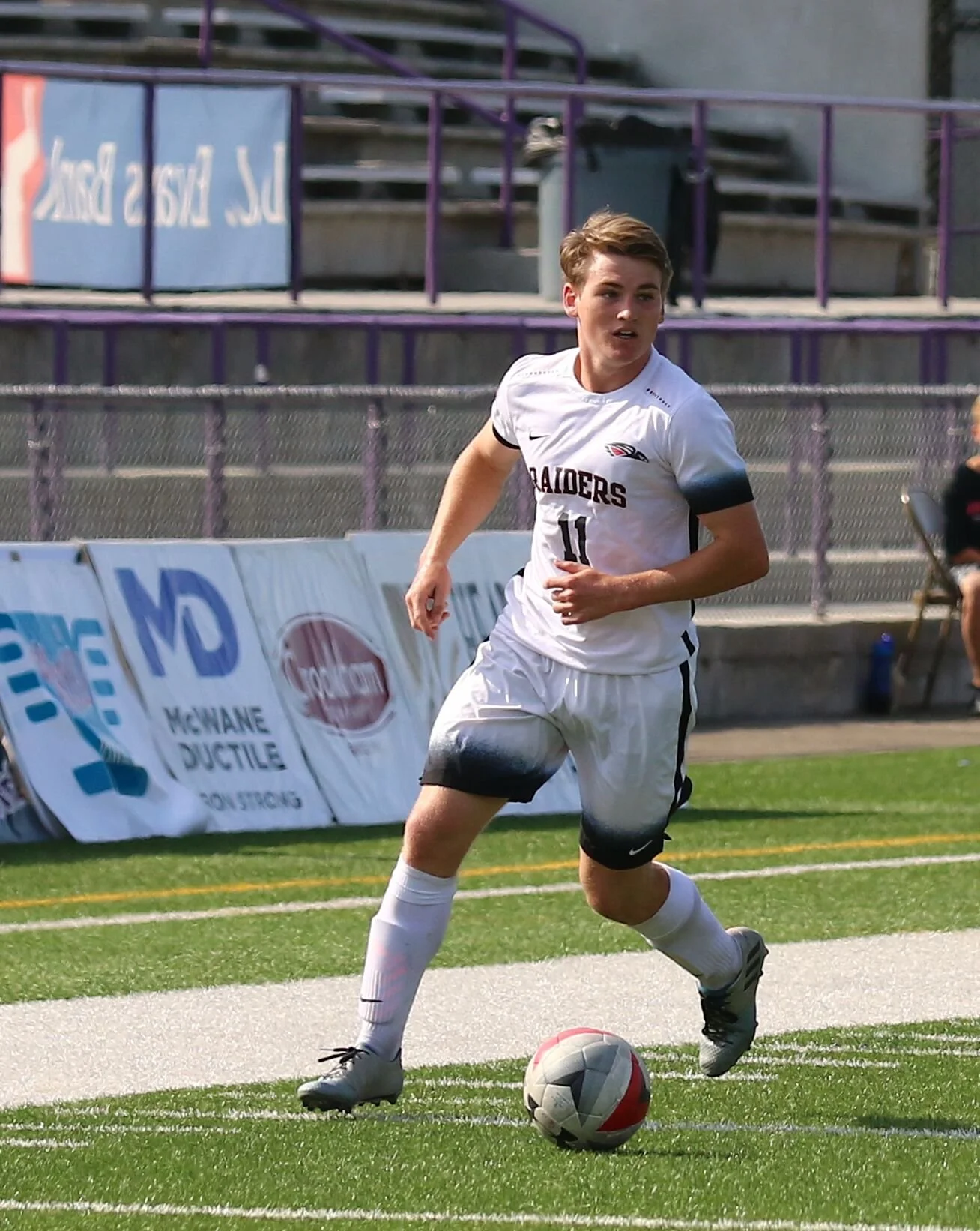 A young male soccer player in a white uniform dribbling a ball on a field.