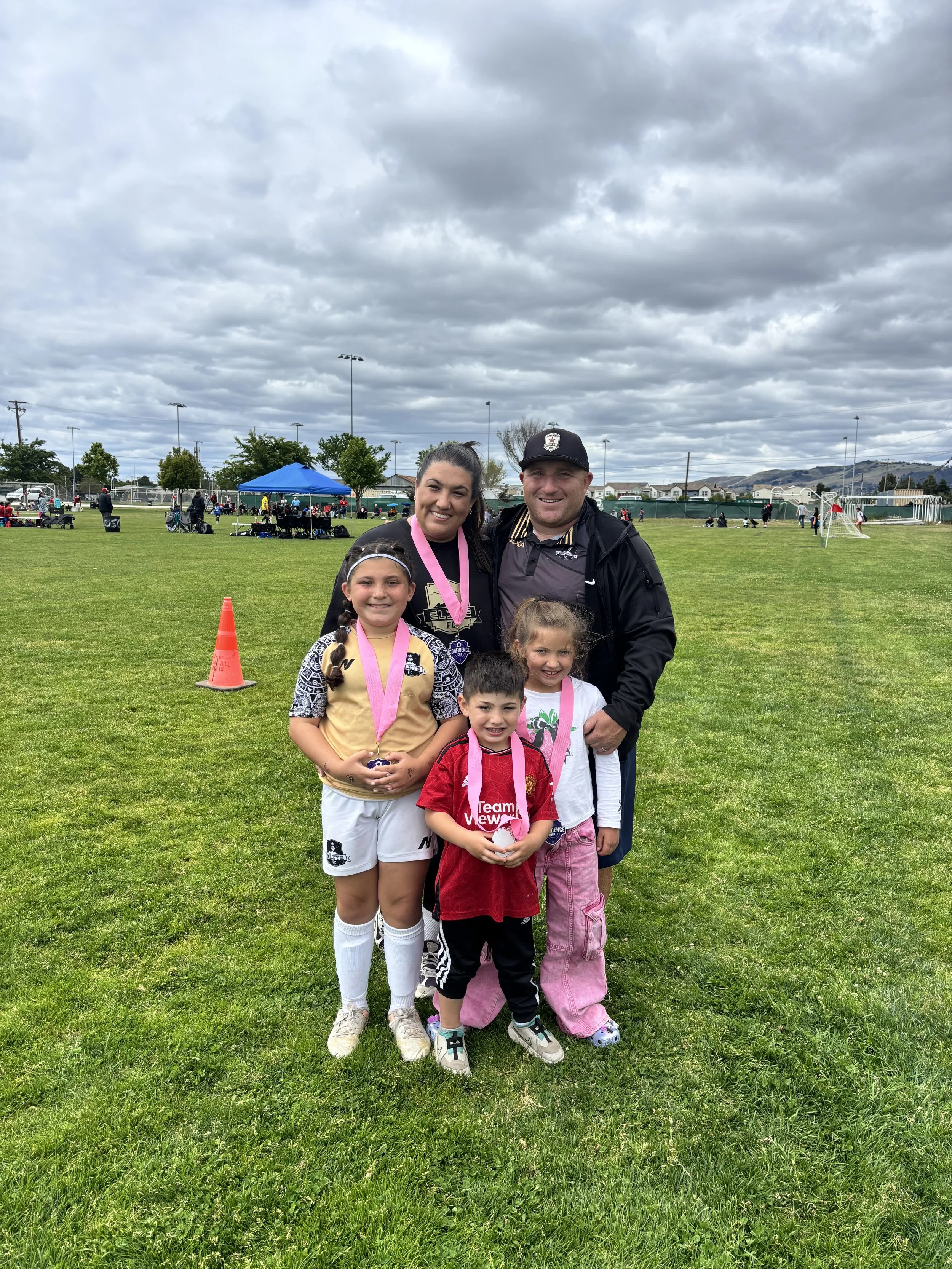 A group of five people, including three children and two adults, stands on a grassy field after a sports event. The children are wearing sports uniforms and medals, smiling at the camera. The background shows a sports field with equipment, tents, and other people, with mostly cloudy skies overhead.