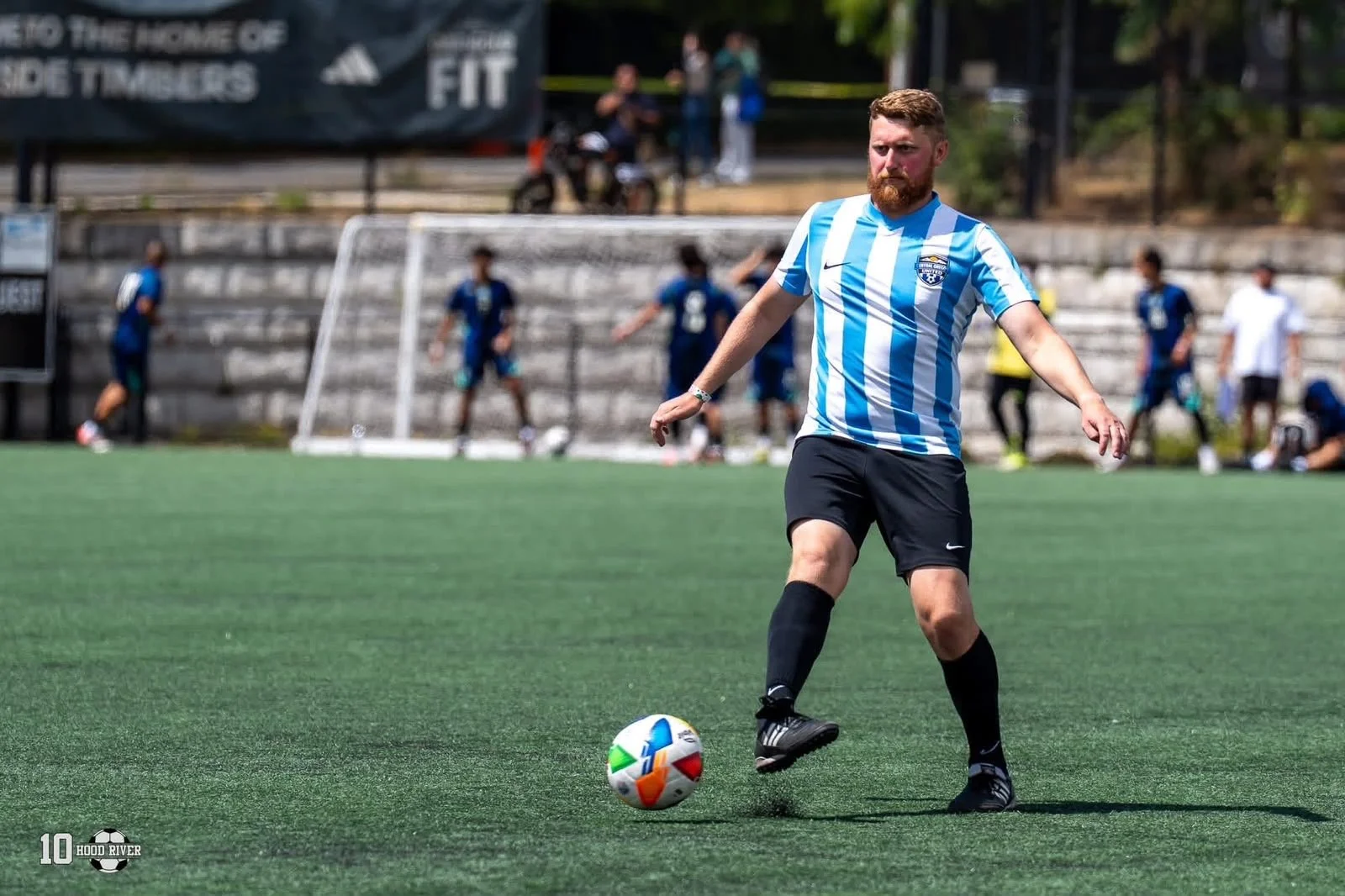 A man with a beard in a blue and white striped soccer jersey and black shorts is about to kick a soccer ball on a green field, with several people and a goal in the background.