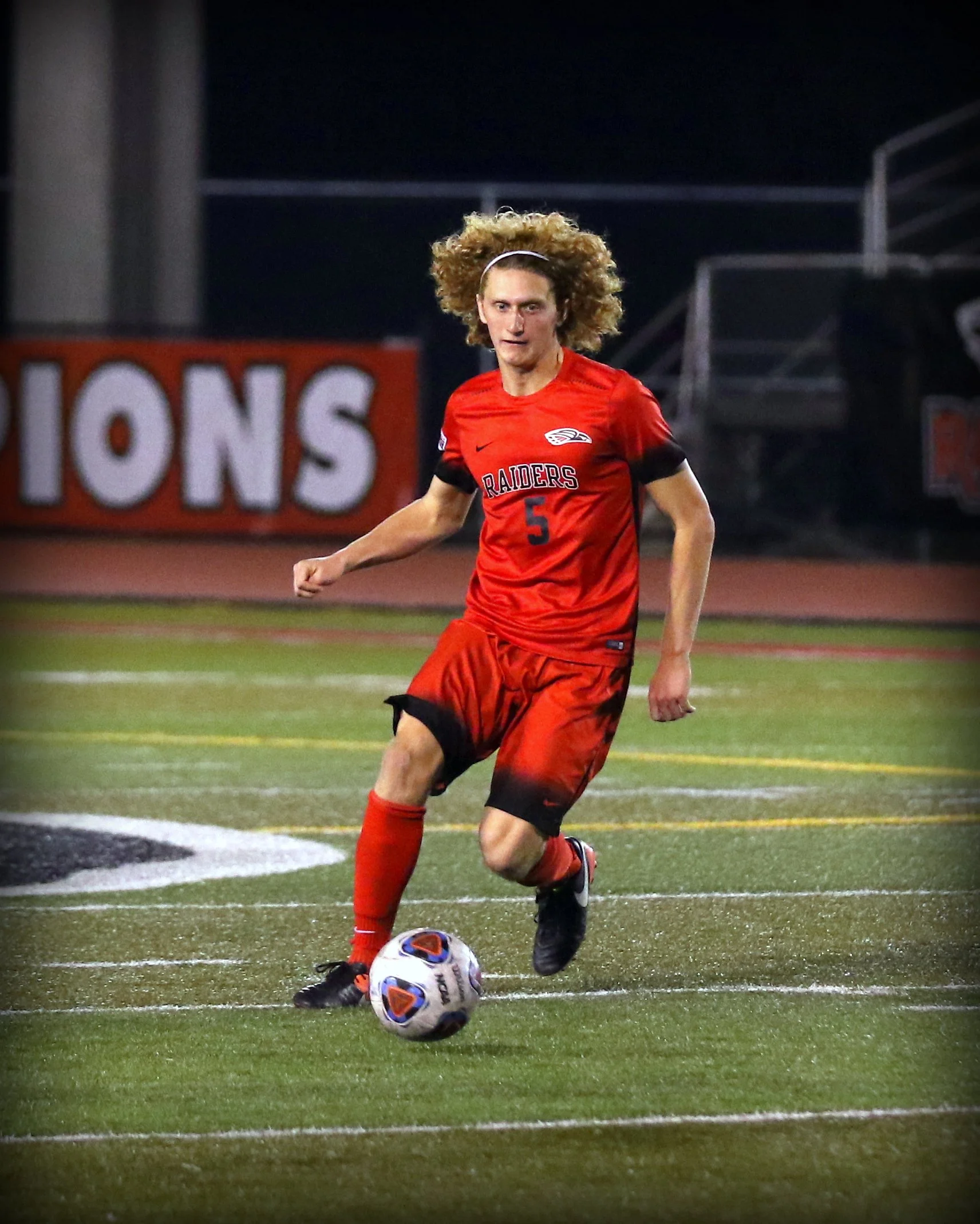 A young male soccer player with curly hair and a headband, wearing a red jersey with the number five, is kicking a soccer ball on a field at night.