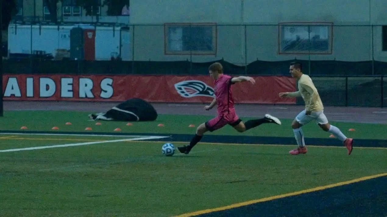 Two soccer players in a match, one in a pink uniform kicking the ball, and another in a yellow uniform running behind.