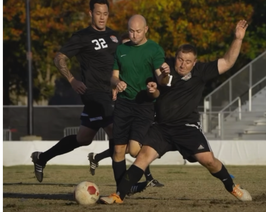 Three men in soccer uniforms competing for the ball on a field during daytime.