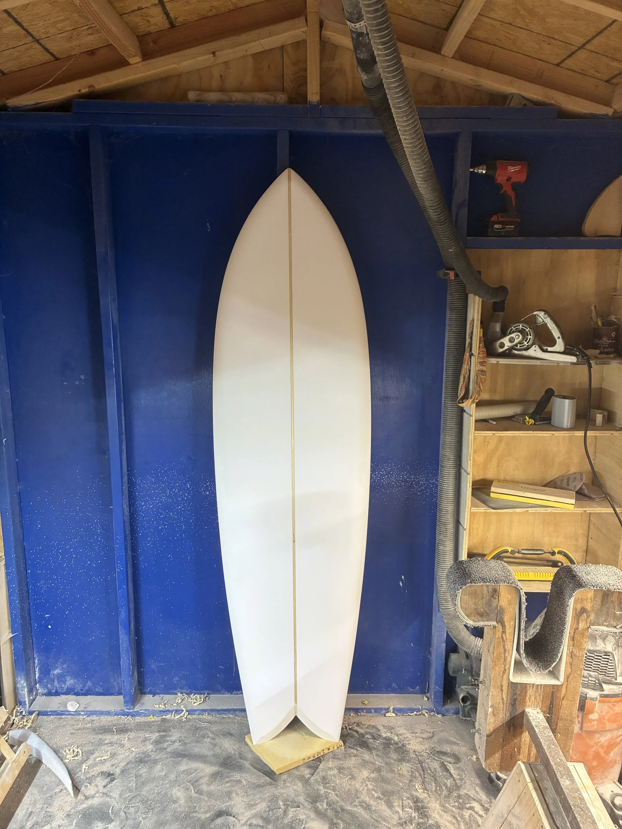 A surfboard is standing upright on a foam block in a woodworking or workshop space. The workshop has a wooden structure with exposed beams, and tools and equipment are visible, including a drill, a sander, and a vacuum hose.