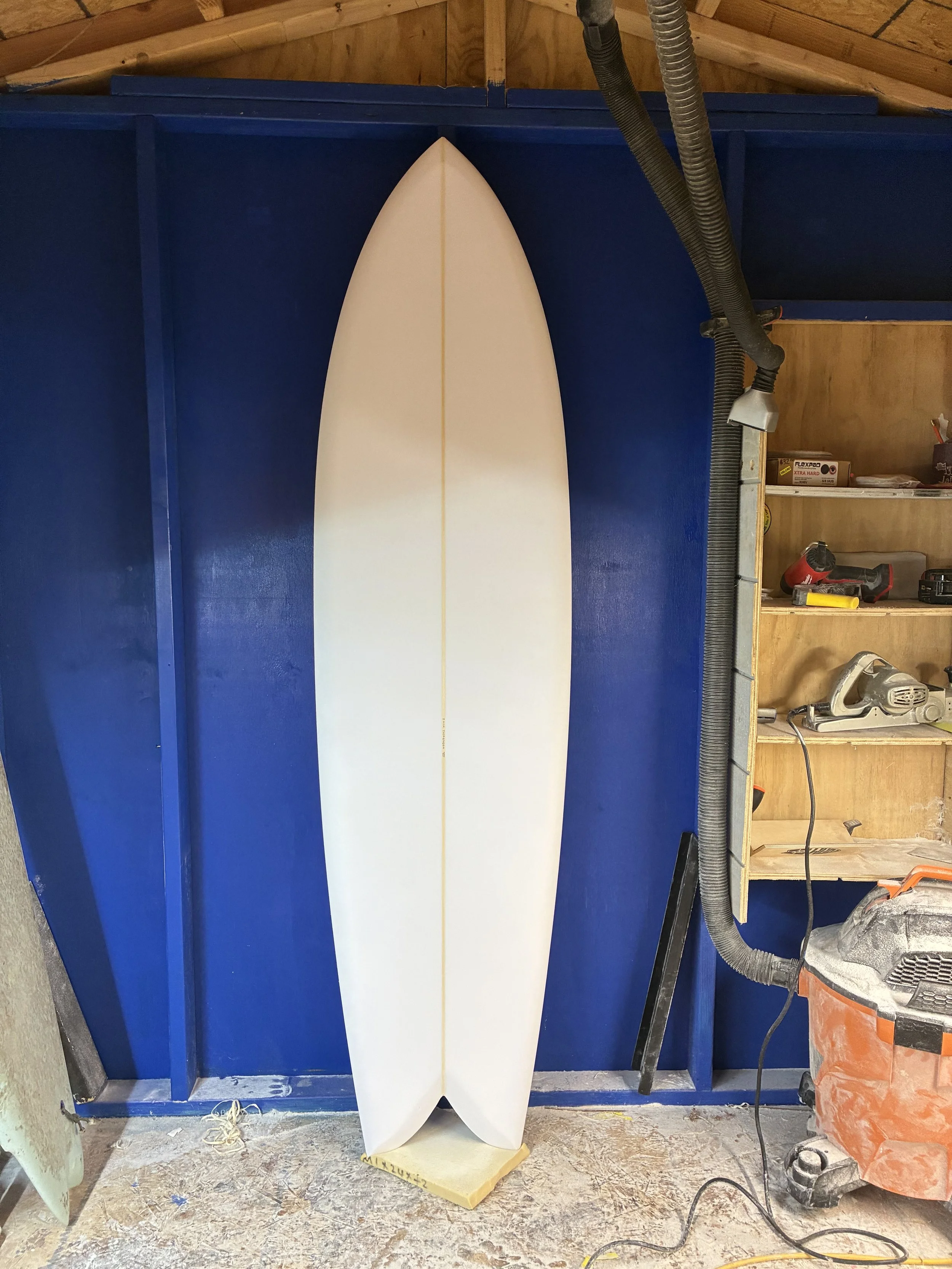A white surfboard leaning against a blue custom-built wall in a workshop with various tools and supplies around.