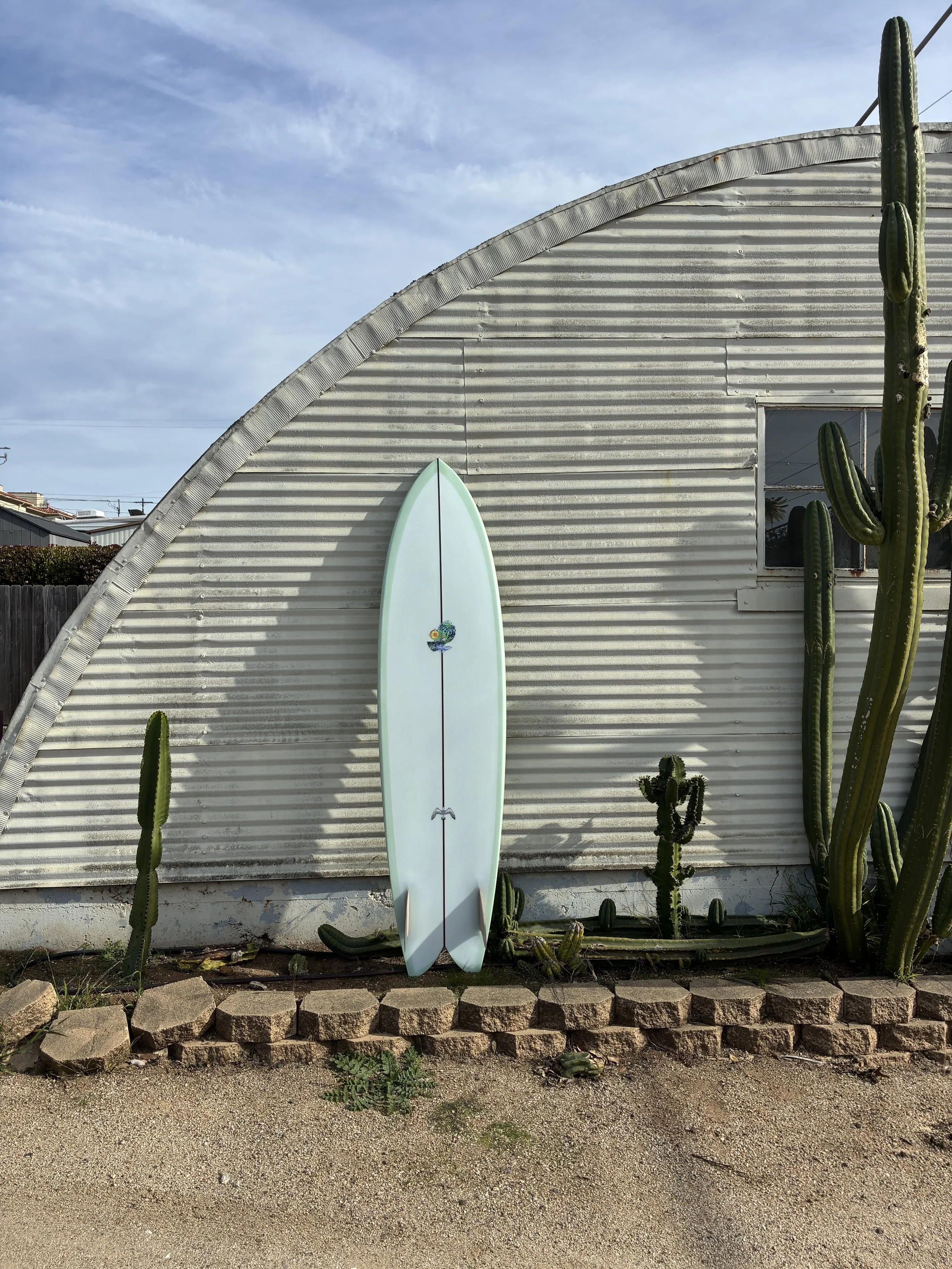 A pale green surfboard with a small graphic near the top, leaning against a corrugated metal building with a window, surrounded by tall cacti, with a dirt and stone border at the bottom, and a partly cloudy sky overhead.