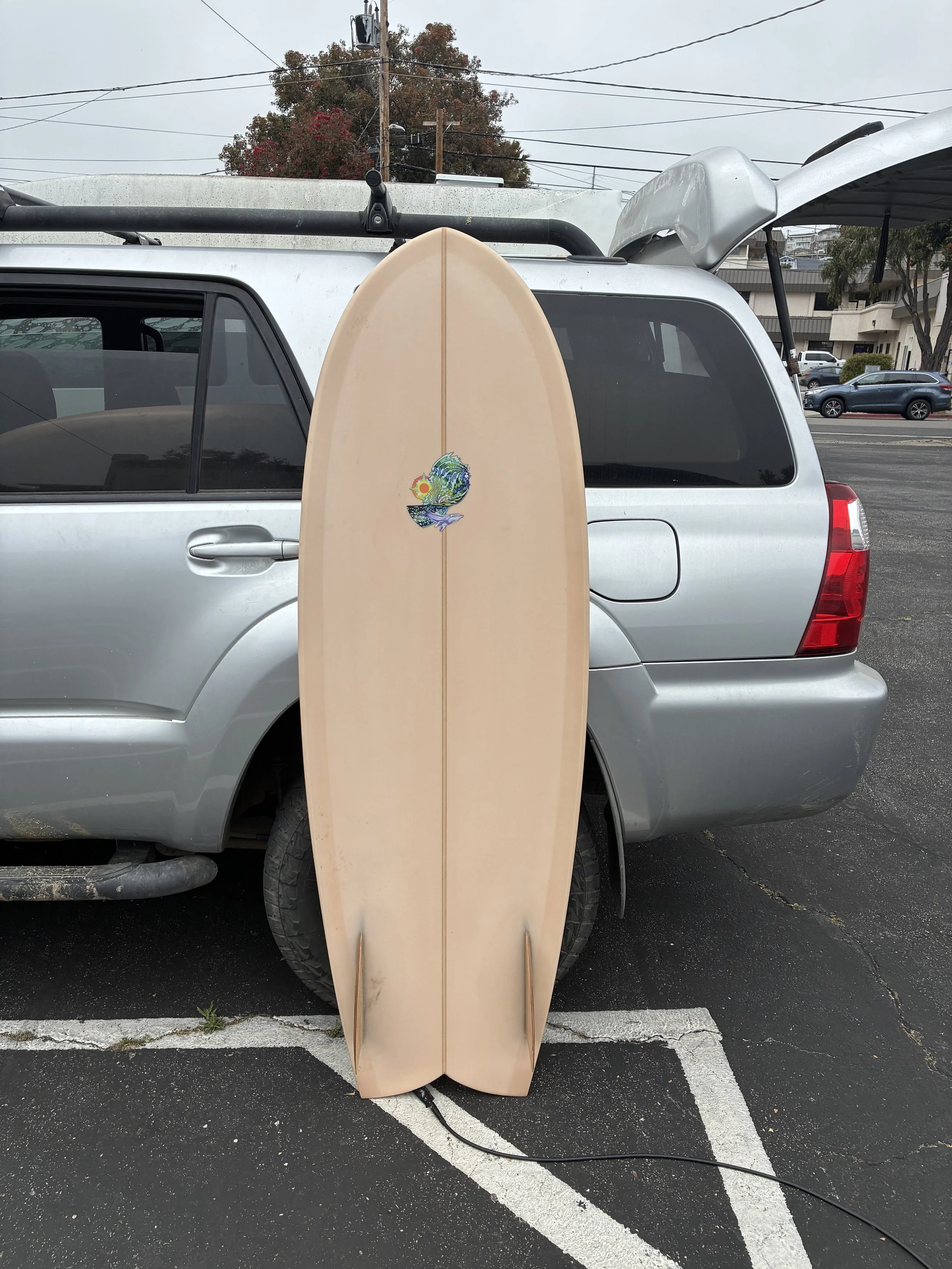 A beige surfboard with a colorful design leaning against the back of a silver minivan in a parking lot.