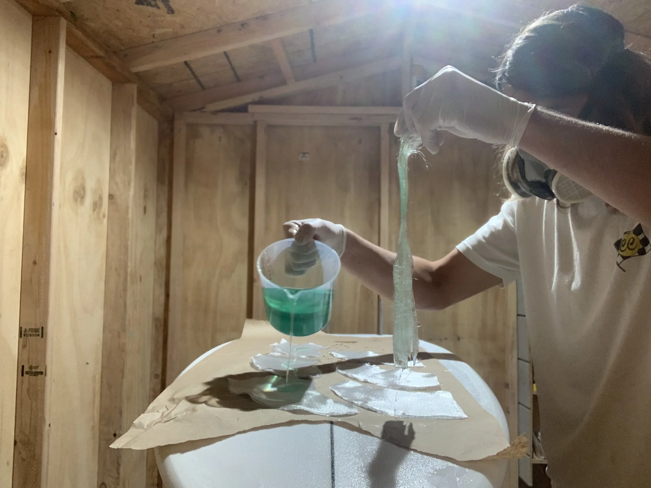 Person pouring a liquid into a green and white cup inside a wooden structure, with sunlight shining in from above.