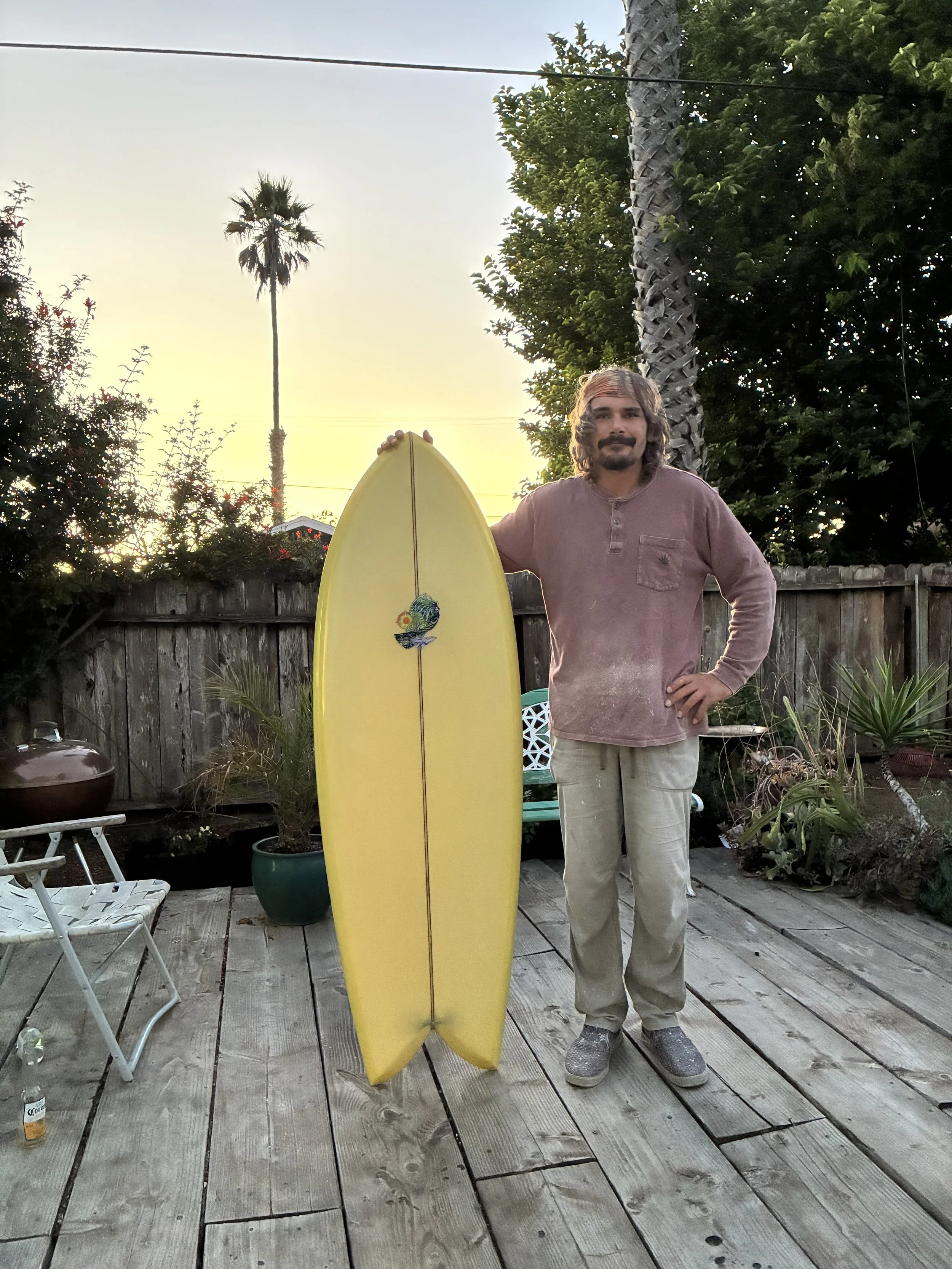 A man with long hair and a beard standing outdoors on a wooden deck holding a yellow surfboard with a colorful logo. He is wearing a mauve long sleeve shirt, beige pants, and gray shoes. The background features a wooden fence, potted plants, trees, and a tall palm tree at sunset.