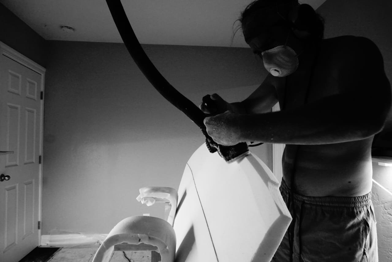 A person wearing a face mask and goggles is sanding or smoothing a surfboard in a garage or workshop.