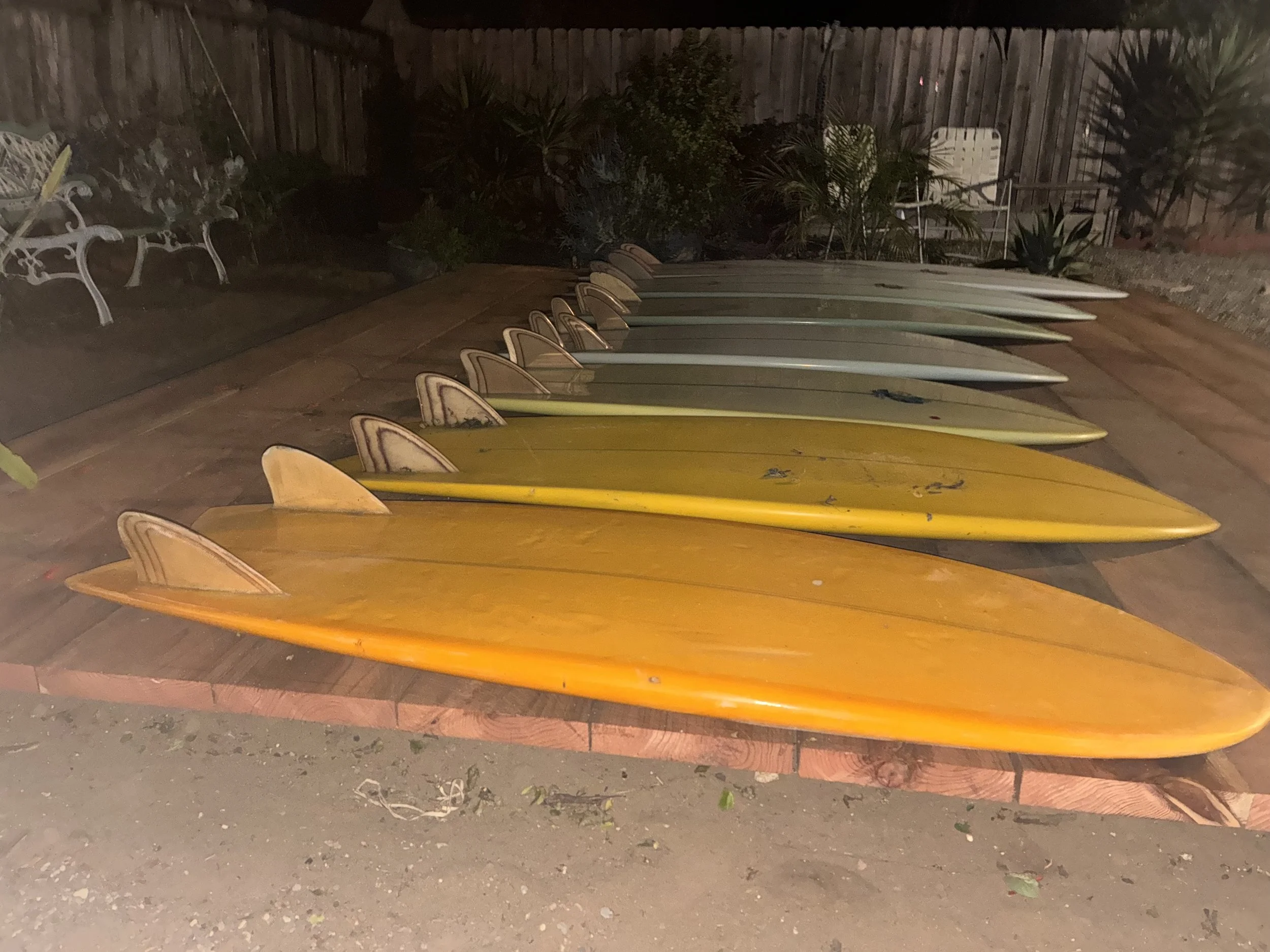 A row of colorful surfboards on the ground, with a garden and wooden fence in the background, during nighttime.