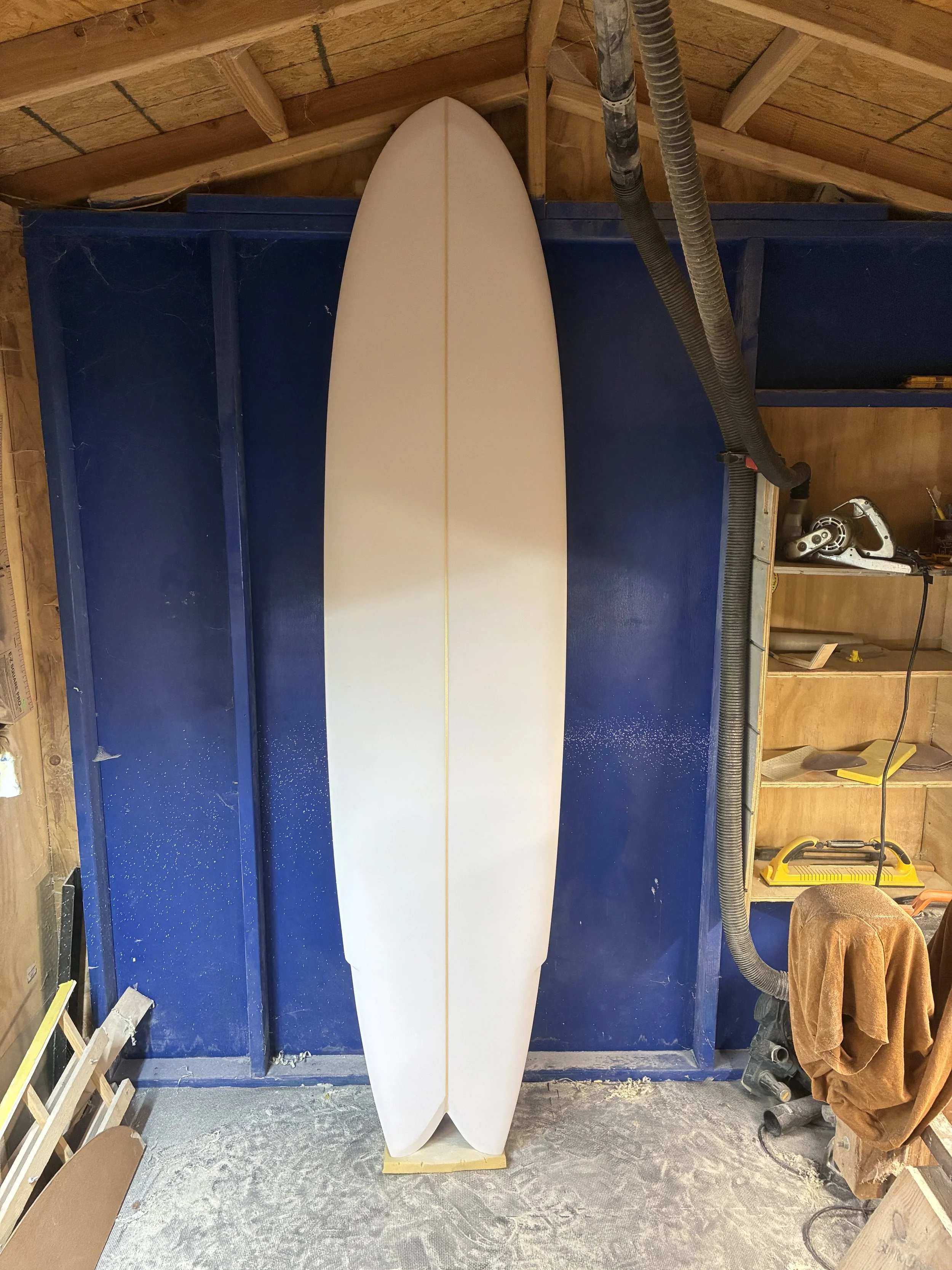 A white surfboard standing upright in a woodworking workshop with tools and wood around.