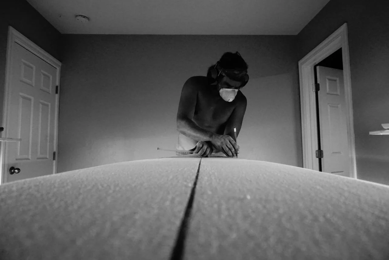 A shirtless man wearing safety goggles and a dust mask stands at a workbench, working on a long piece of material, in a room with open doorways.