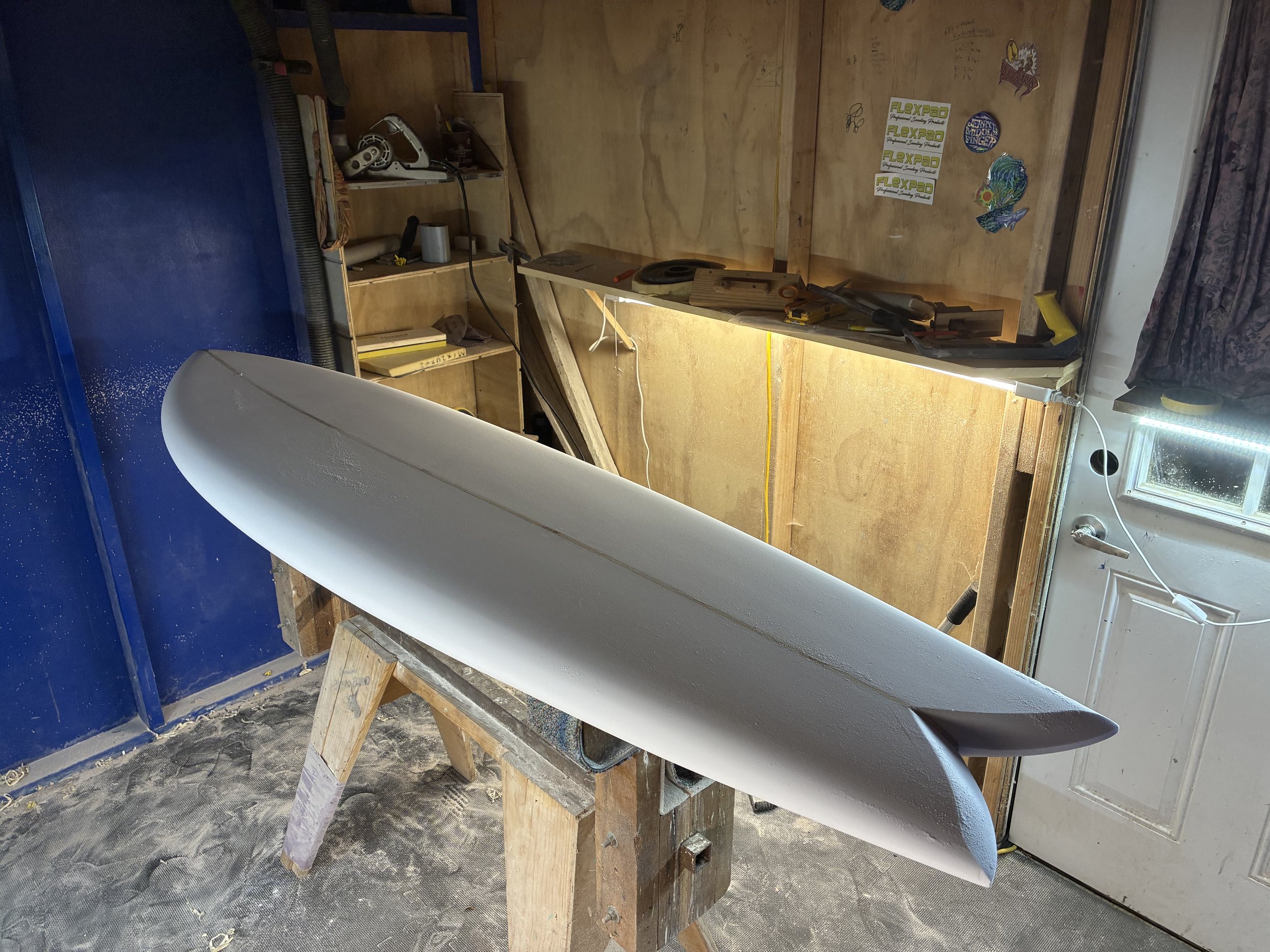 A workspace with a surfboard mounted on a makeshift wooden stand inside a room with plywood walls. The workspace has various tools and items, including a drill, yellow tape, and a small wooden box. Stickers and decals are on the wall, and there's a window with a floral curtain. The floor appears dusty and is likely a garage or workshop.