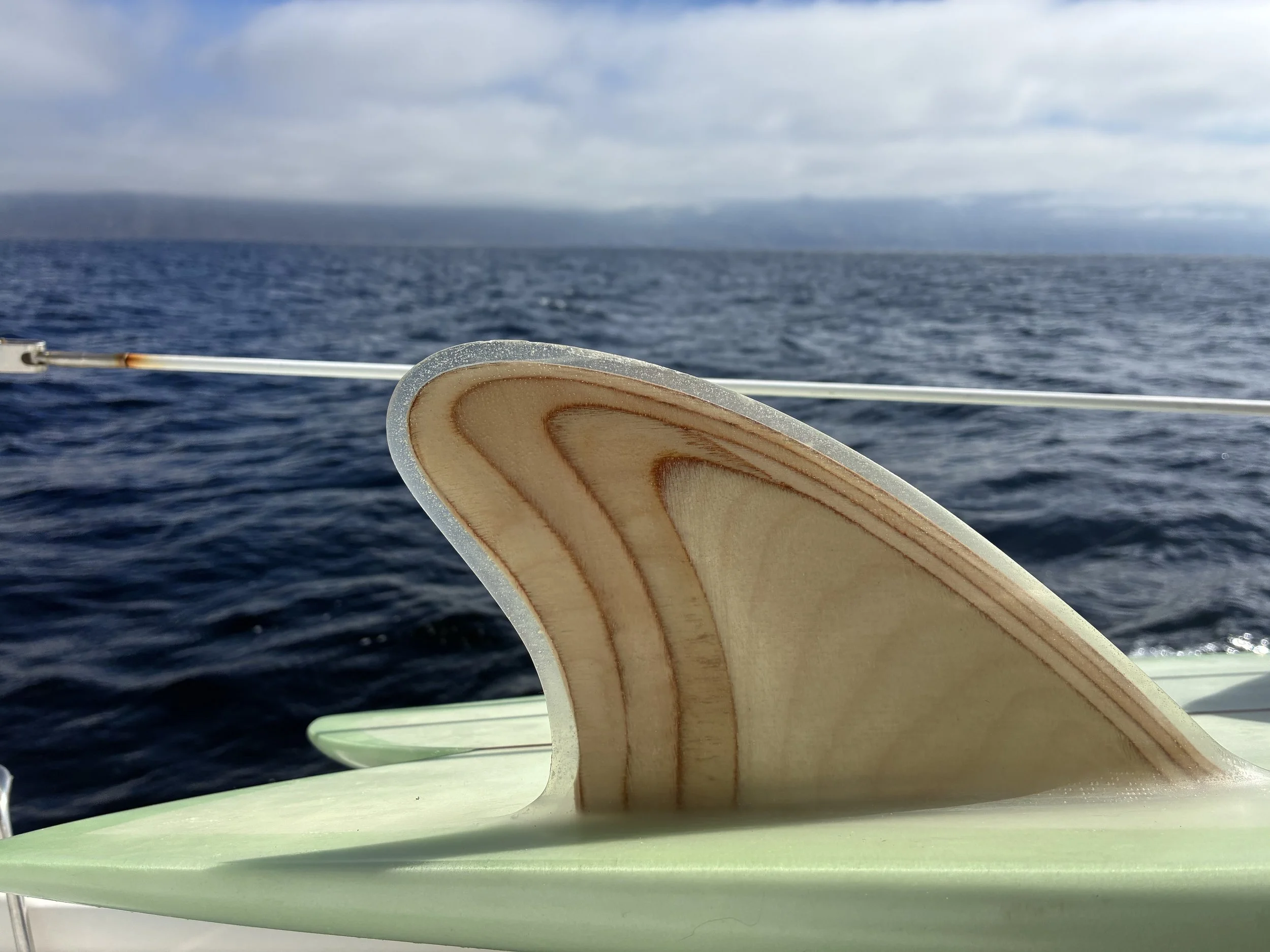 Close-up of a wooden surfboard fin used on a surfboard, with ocean and cloudy sky in the background.