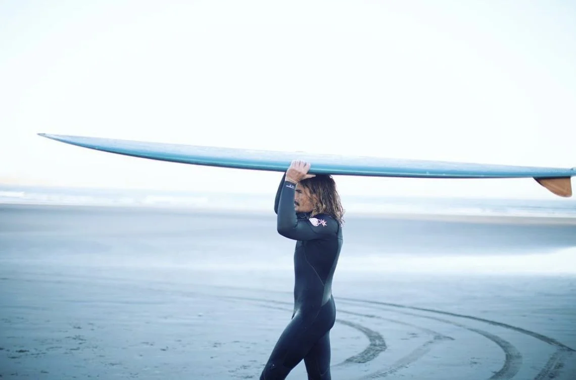 Person in wetsuit carrying a surfboard on a sandy beach.