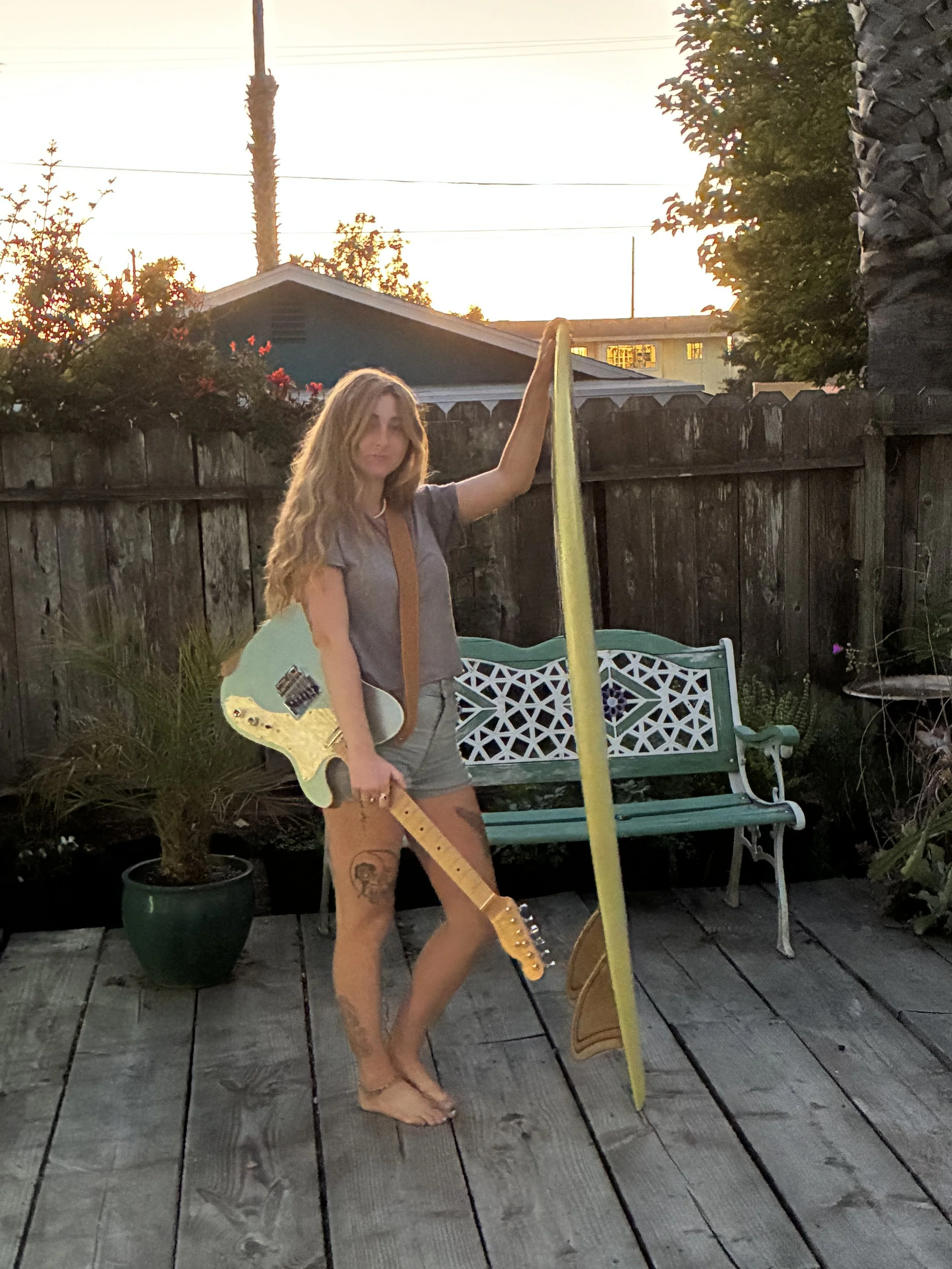 A woman with long, wavy blonde hair, wearing shorts and a t-shirt, standing outdoors on a wooden deck at sunset. She is holding a yellow guitar and leaning against a slide, with a wooden fence, some potted plants, a bench, and trees in the background.