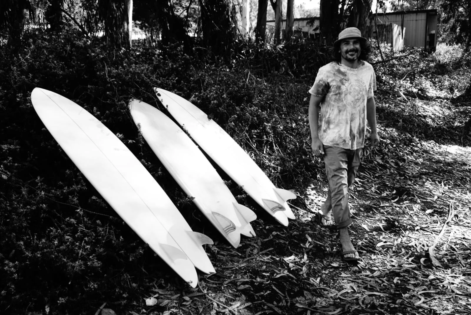 A man with long hair and a beard, wearing a hat, a tie-dye t-shirt, and flip-flops, walking on a dirt path with three surfboards leaned against bushes to his left. The background shows foliage and trees.