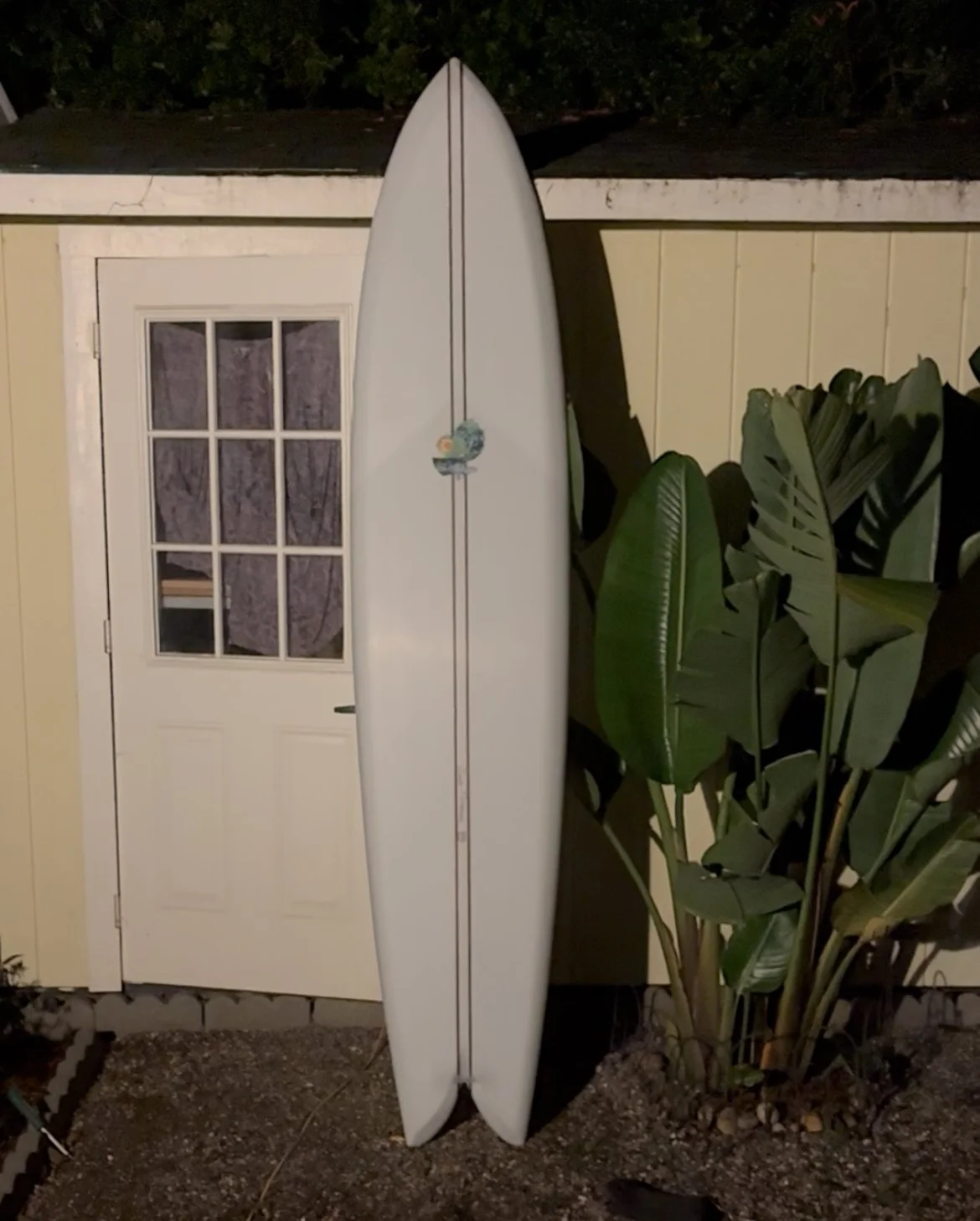 A white surfboard leaning against a yellow wall outside a building, with large green leaves nearby at night.