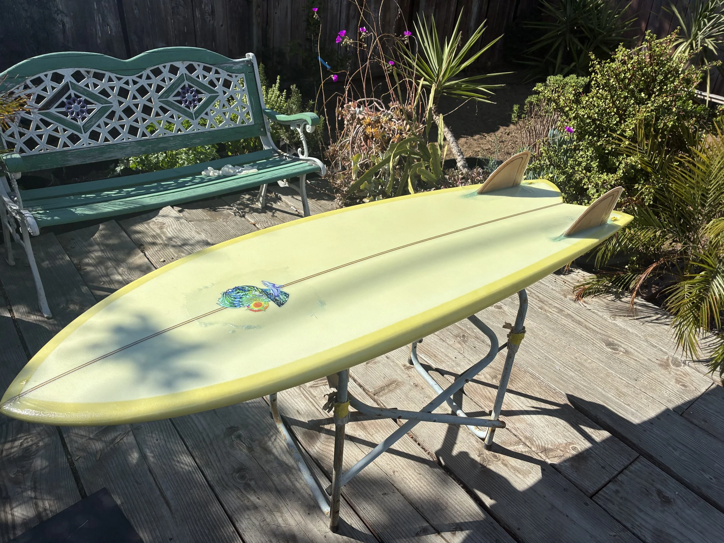 A backyard patio with a green decorative metal bench on a wooden deck, surrounded by tropical plants and flowers, with sunlight casting shadows.