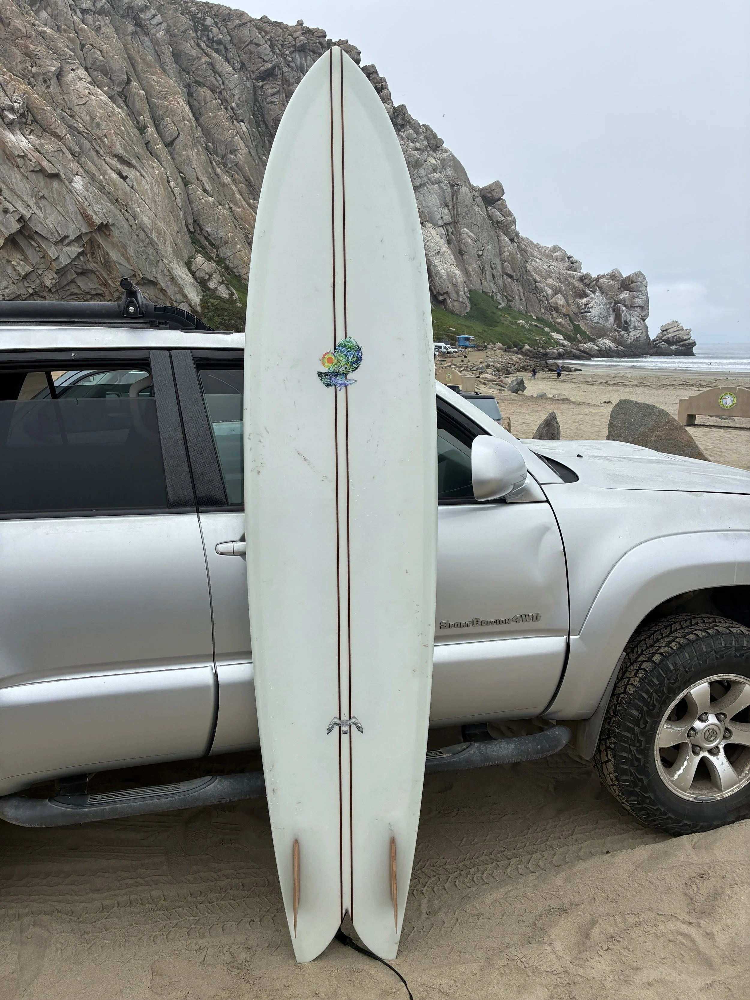 A white surfboard with a colorful wave design and a tail fin, leaning against a silver pickup truck on a sandy beach with large rocks and cliffs in the background.