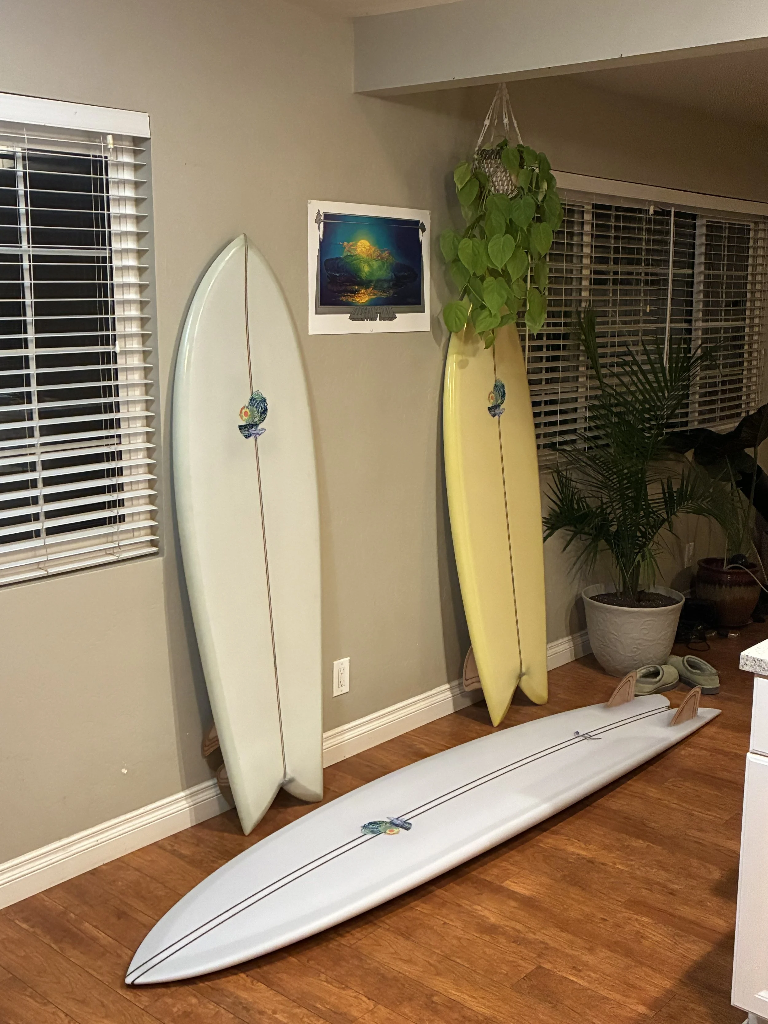 Two surfboards, one white and one yellow, leaning against a wall in a room with wood flooring. There is a potted plant, a window with blinds, and a picture on the wall.