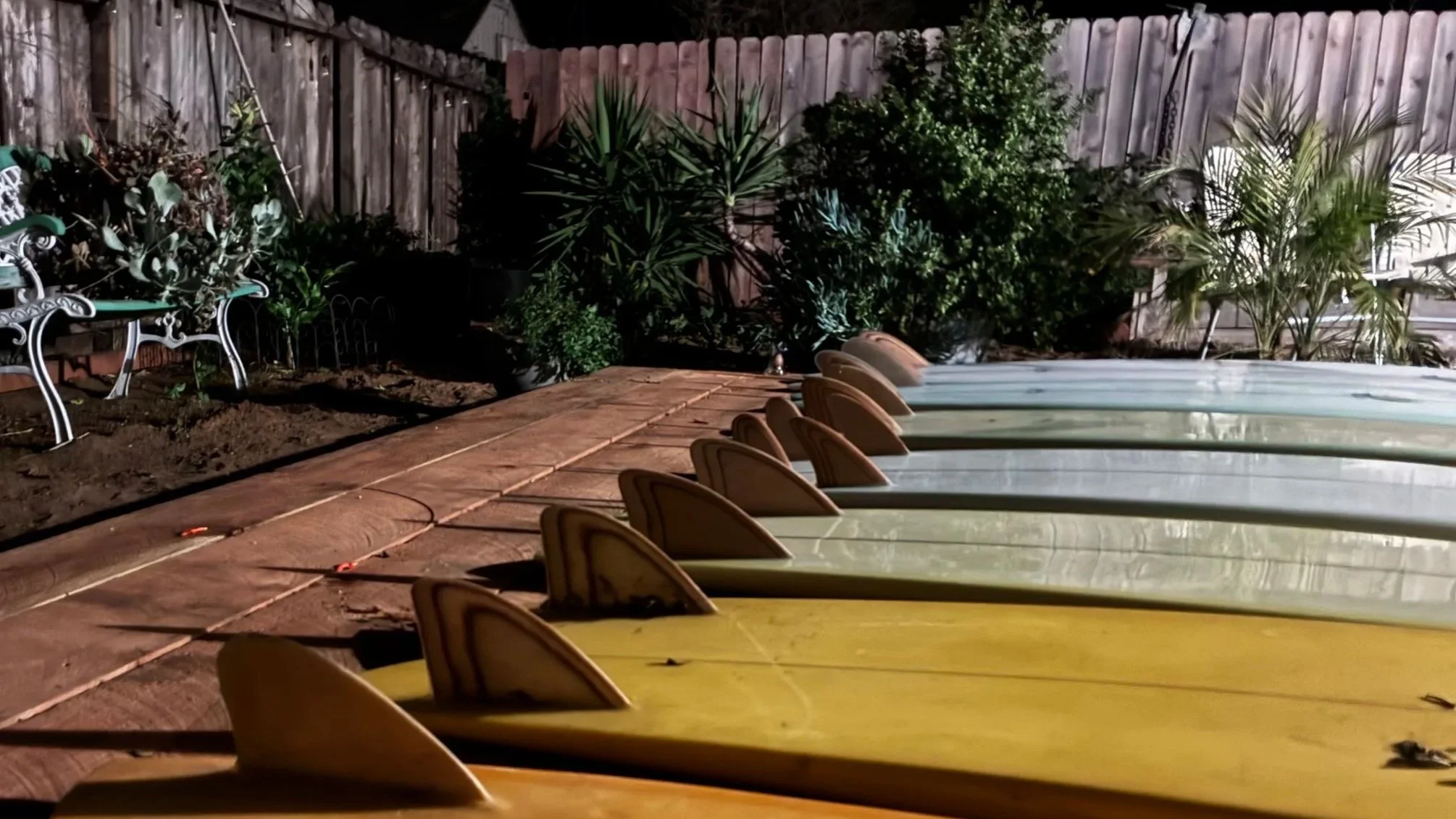 A backyard patio at night with a row of empty plastic lounge chairs in light green, yellow, and orange, lined up along the perimeter. Behind the chairs, there is a garden with various green plants, including a palm-like plant and a bush, with a wooden fence in the background. A metal garden chair with a decorative backrest is on the left side. The yard is lit by an outdoor light, creating shadows on the ground and fence.