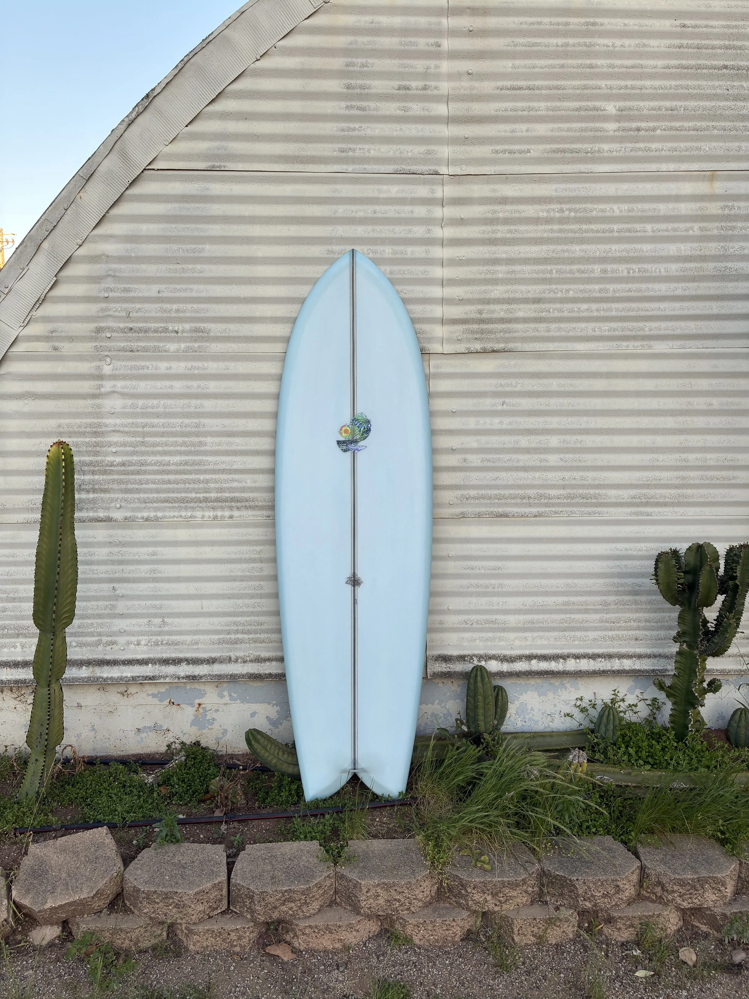 A light blue surfboard with a decorative sticker stands upright against a white, textured metal building. Two tall cacti are on either side of the surfboard, and smaller plants and rocks are lined at the base.