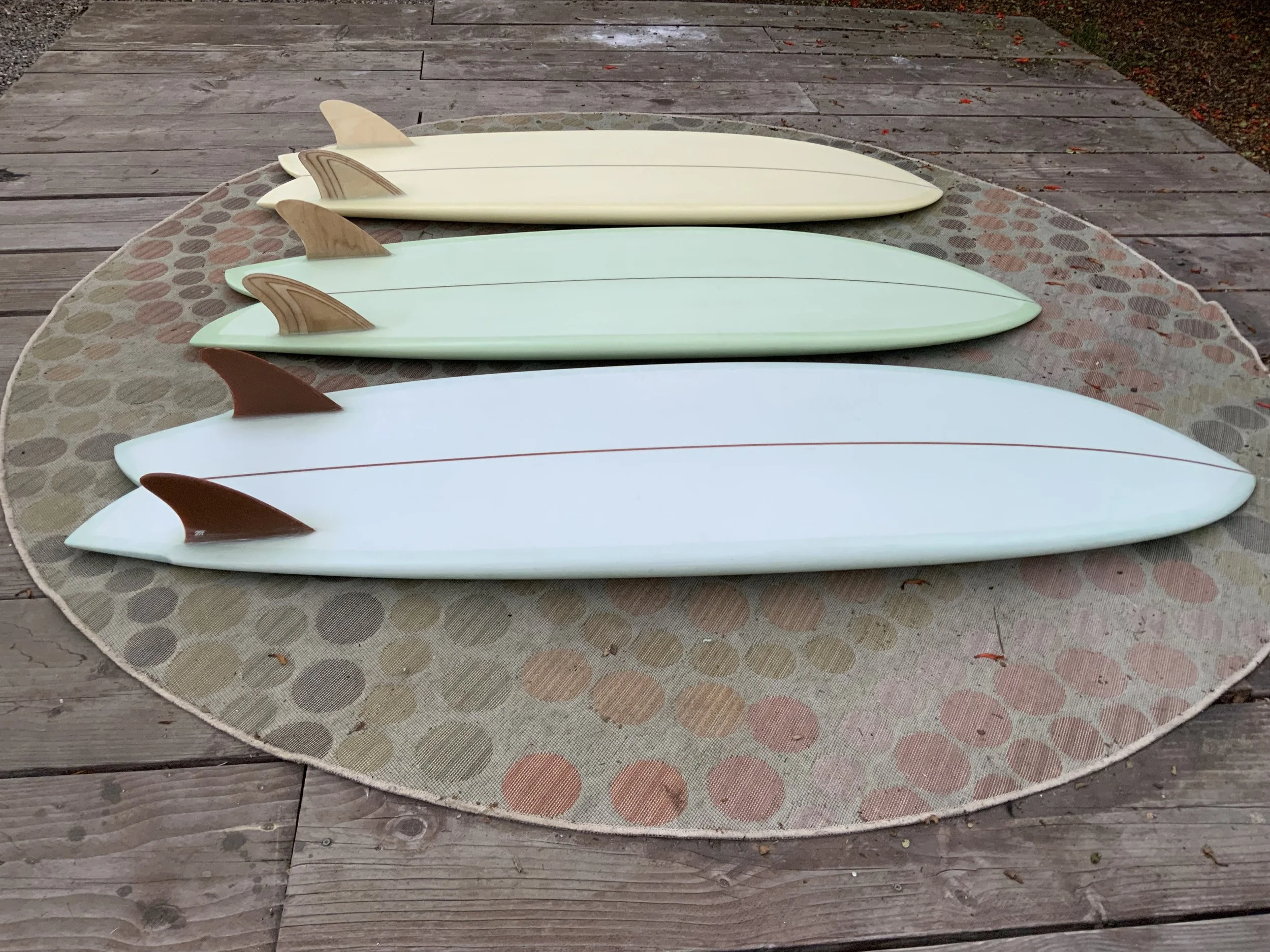 Four surfboards with dorsal fins laid out on a round picnic table with a patterned tablecloth, on a wooden deck outdoors.