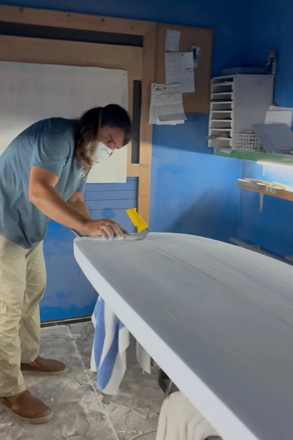 A person in a blue shirt and khaki pants sanding a surfboard in a workshop with blue walls and shelves.