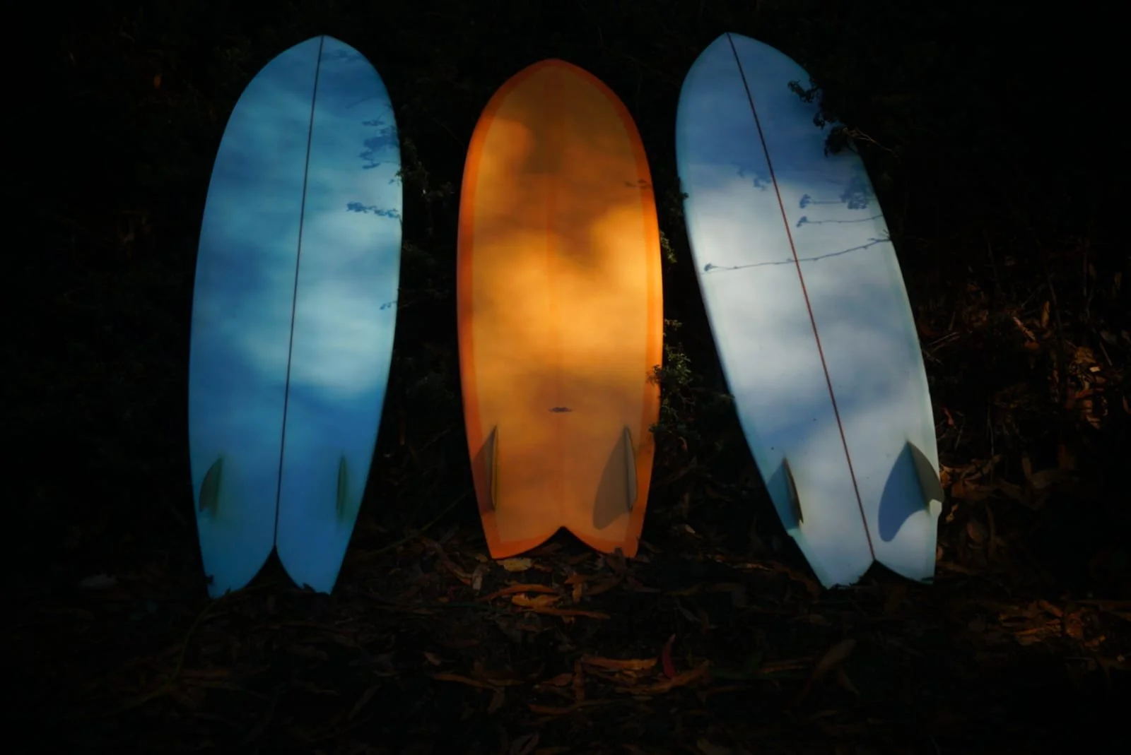 Three surfboards strung up against a dark background, illuminated from below. The left and right surfboards are blue with cloud patterns, and the middle surfboard is orange.