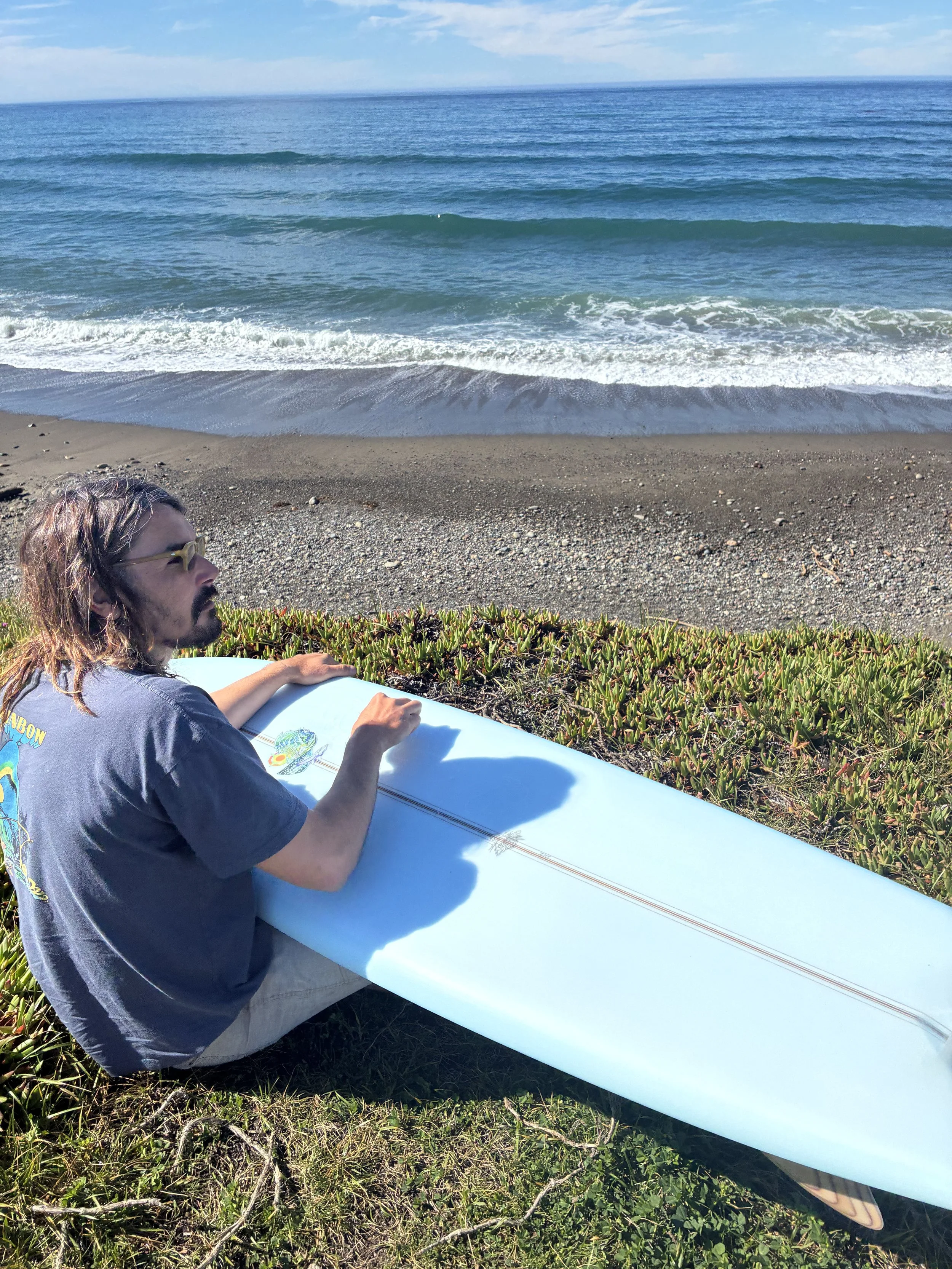 A man with long hair and glasses sitting on a grassy area holding a white surfboard near the beach with ocean waves and a blue sky in the background.