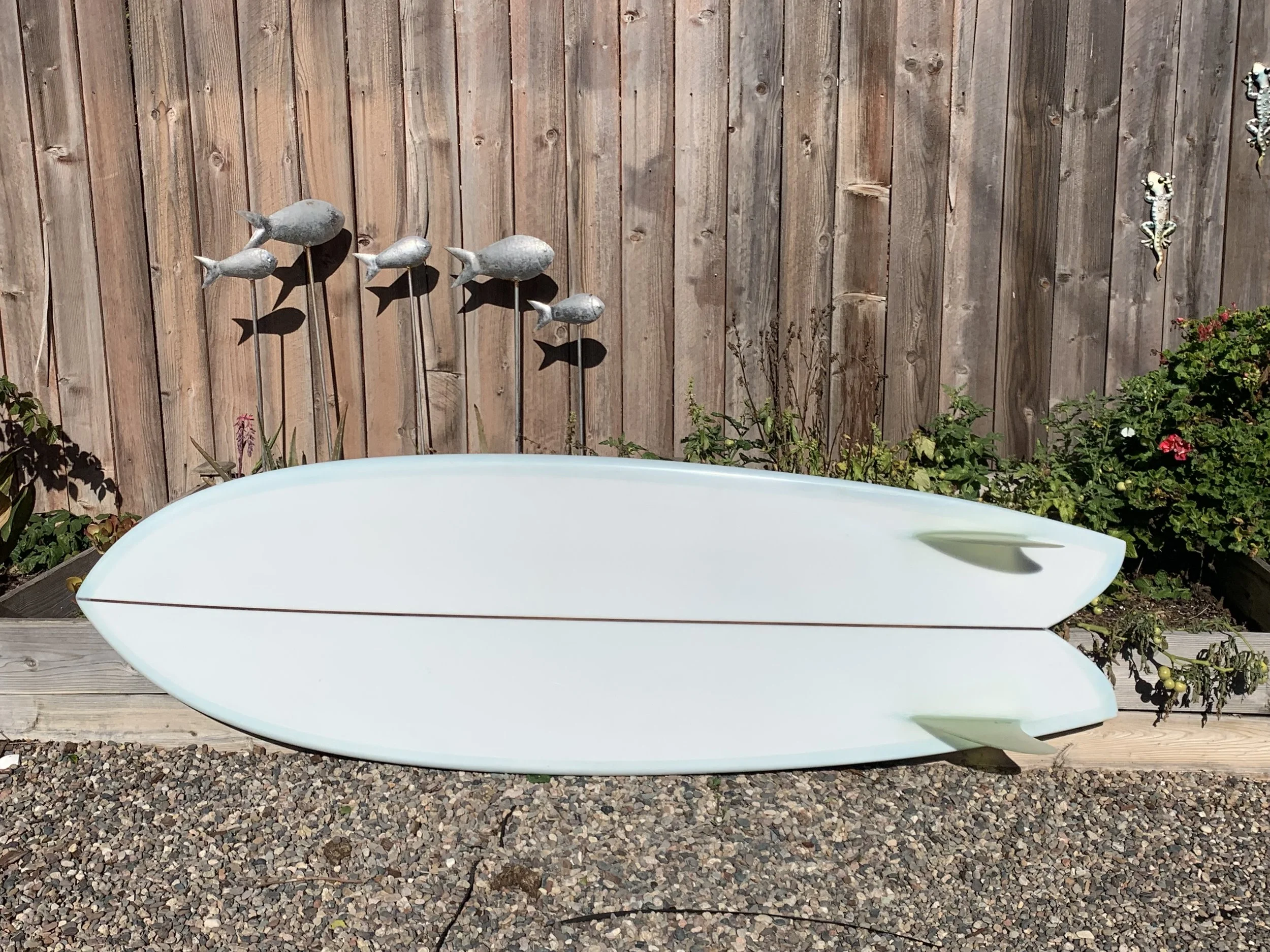 Decorative garden scene with a row of four gray fish-shaped garden ornaments mounted on sticks, a white boat-shaped planter, and a wooden fence with a gecko decoration on the right side, surrounded by various plants and flowers.