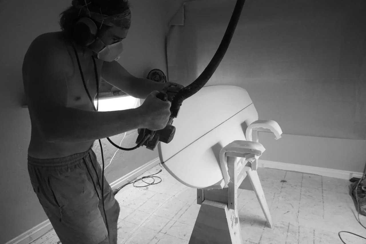 A person in a mask and headphones is using a power tool to carve or shape a white surfboard mounted on a wooden stand in a workshop.