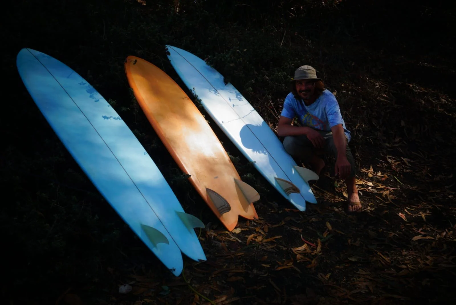 Man crouching next to four surfboards lying on the ground in a forested area during dusk or dawn.