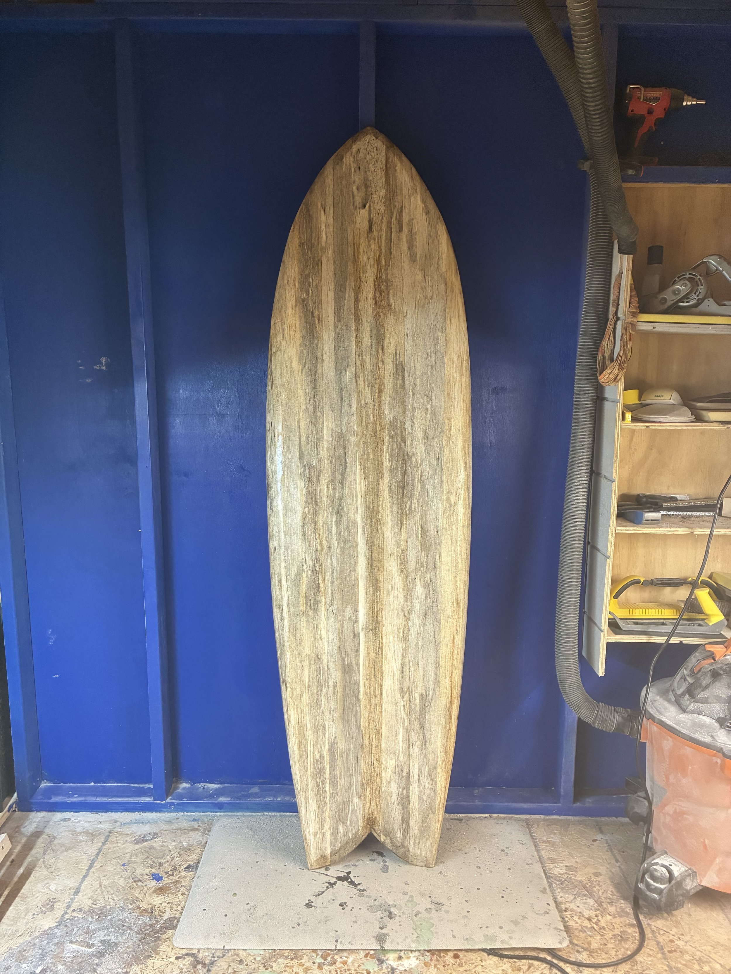 A wooden surfboard with a pointed nose and rounded tail, standing upright in a workshop against a blue wall.