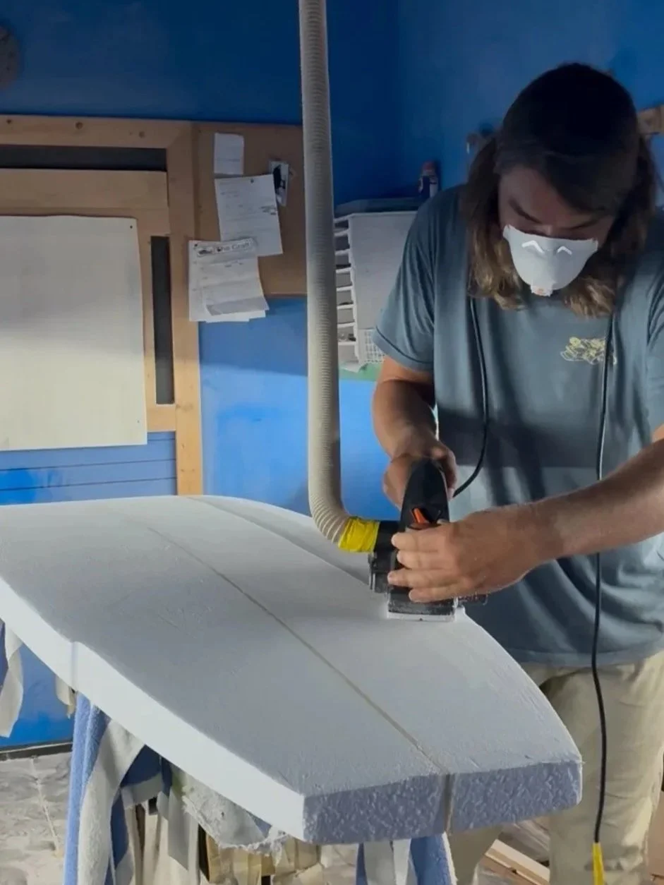 Person wearing a mask and gray shirt is shaping a surfboard foam blank with a hand planer in a workspace.