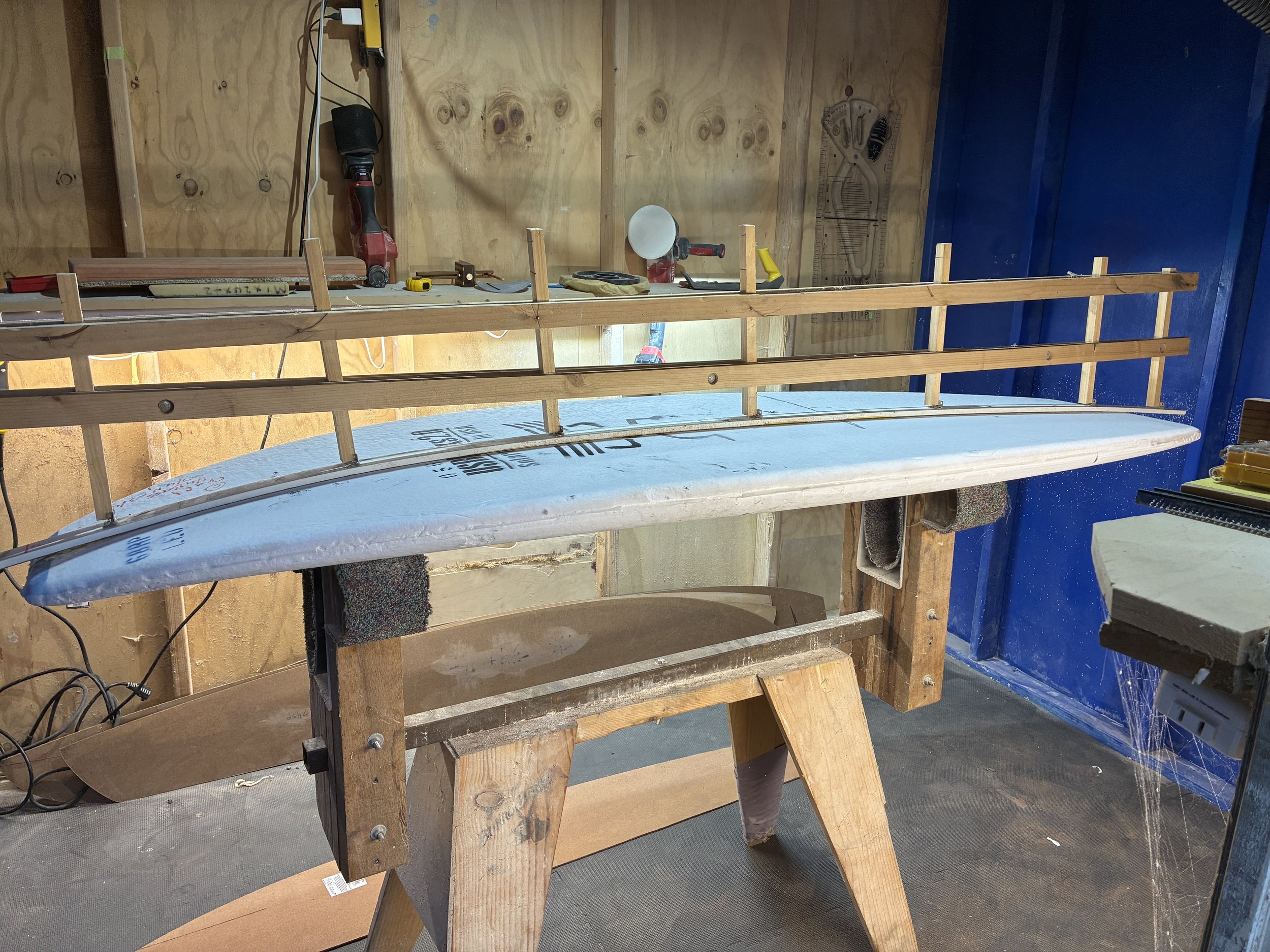 Workshop with a surfboard propped up on wooden supports, surrounded by tools and equipment on a workbench.