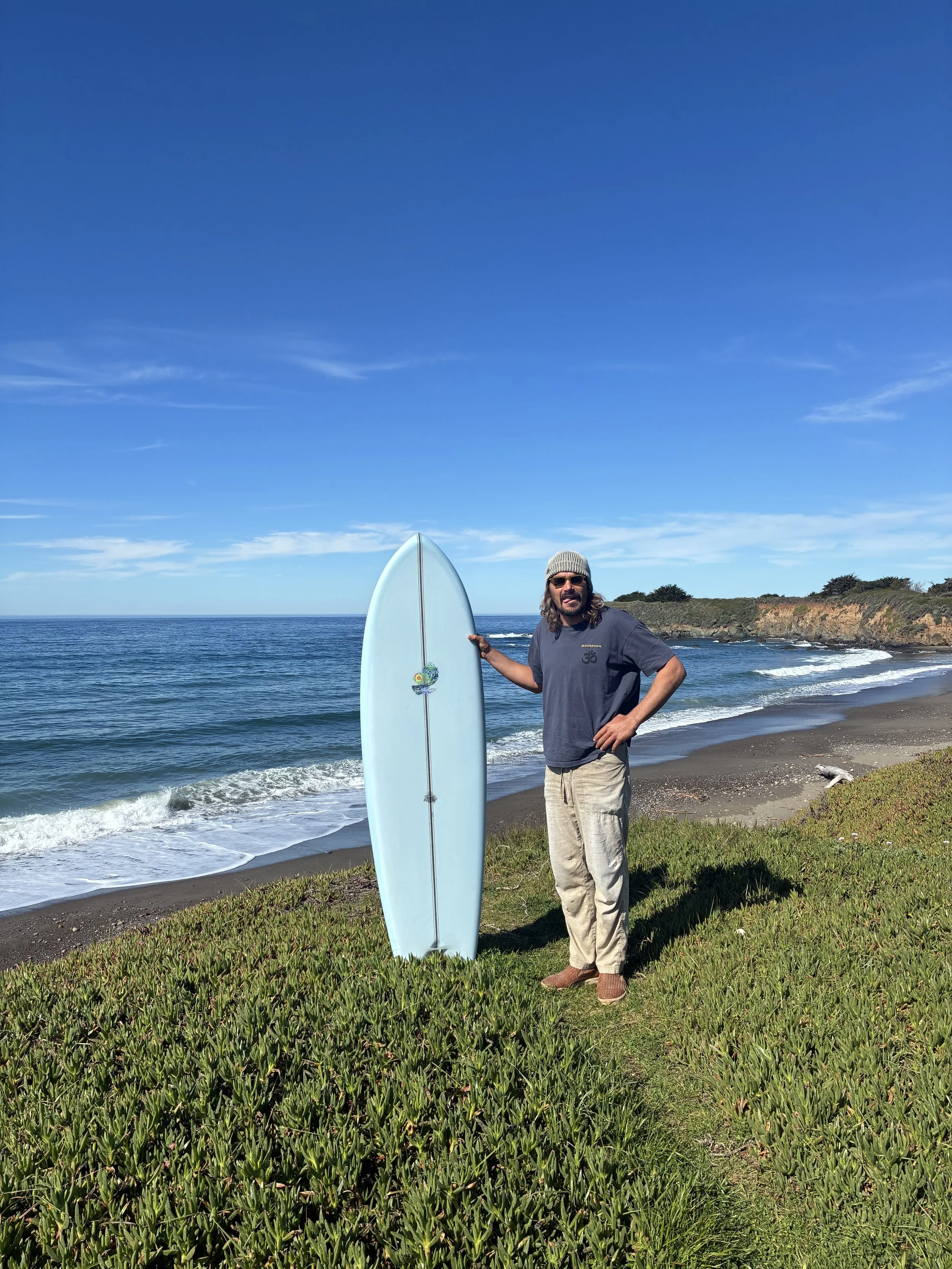 A man with long hair, wearing sunglasses, a knit beanie, a gray t-shirt, and beige pants, stands on a grassy area at the beach, holding a white surfboard upright near the shore with the ocean and cliffs in the background under a blue sky.
