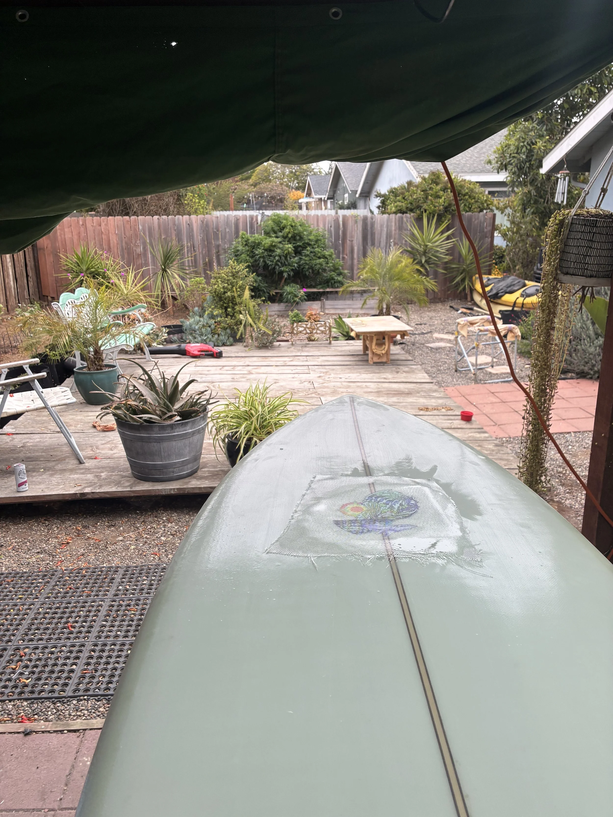 View of a backyard with potted plants, a wooden deck, a small table, a garden fence, and houses in the background. Taken from underneath a canopied or covered area, with an umbrella or tarp above.