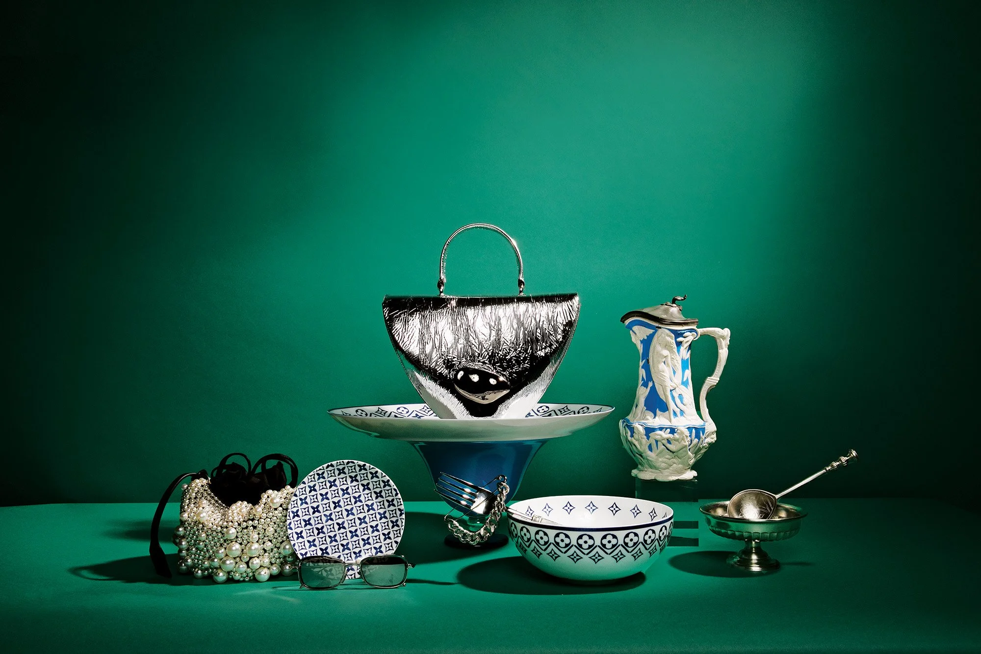 Arrangement of jewelry and tableware on a green background, including pearl necklaces, a pair of eyeglasses, patterned bowls, and a silver spoon.