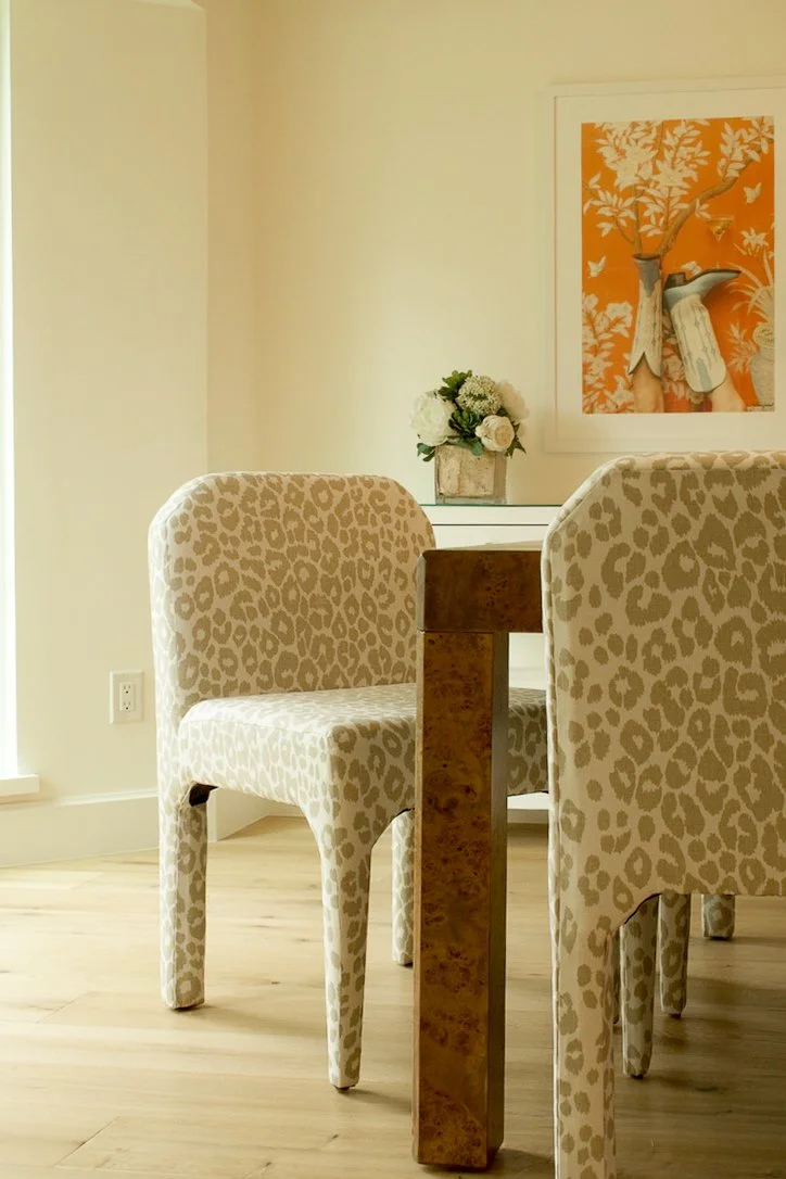 A cozy dining area with a wooden table, two leopard print upholstered chairs, a white sideboard with a floral arrangement, and wall art featuring a woman's hands holding a branch in front of an orange background with butterflies.