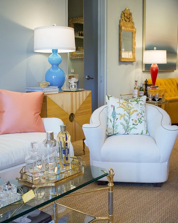 Living room with a white sofa, pink and floral pillows, a glass coffee table with liquor bottles and glasses, a blue table lamp, a wooden side table, and a yellow chair in the background, with decorative mirrors on the wall.