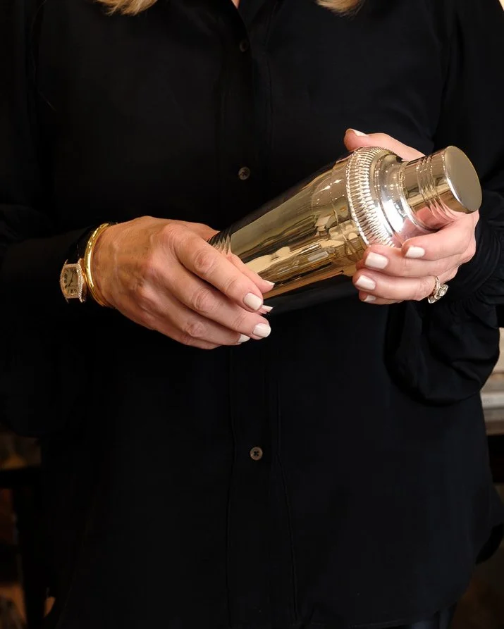 Person dressed in black holding a shiny gold flask with both hands, wearing a gold bracelet and a ring.