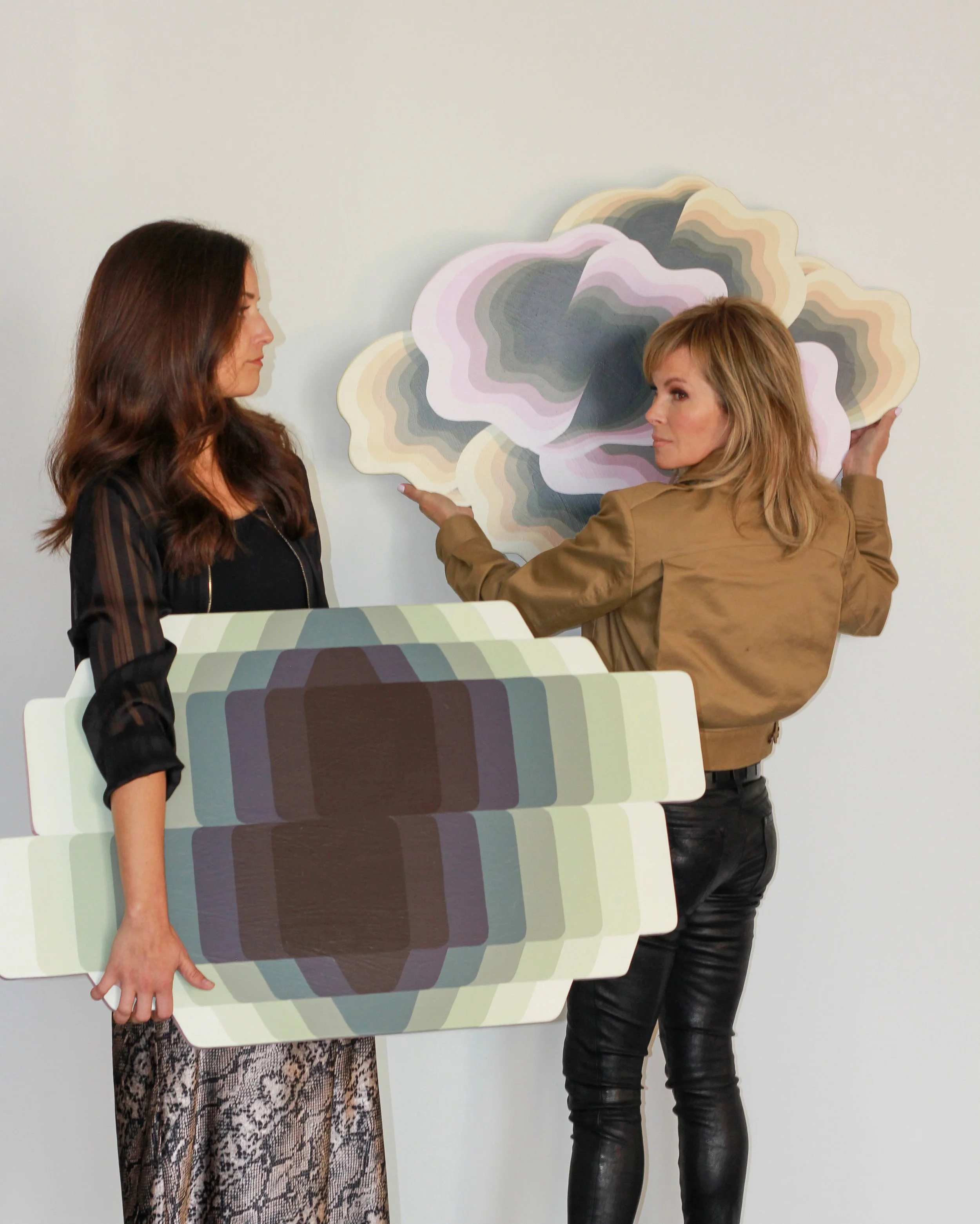 Two women are involved in an activity with large color swatches. One woman, with long dark hair and wearing a black top, holds some color sample cards in one hand. The other woman, with shorter blonde hair and dressed in a tan leather jacket, is adju