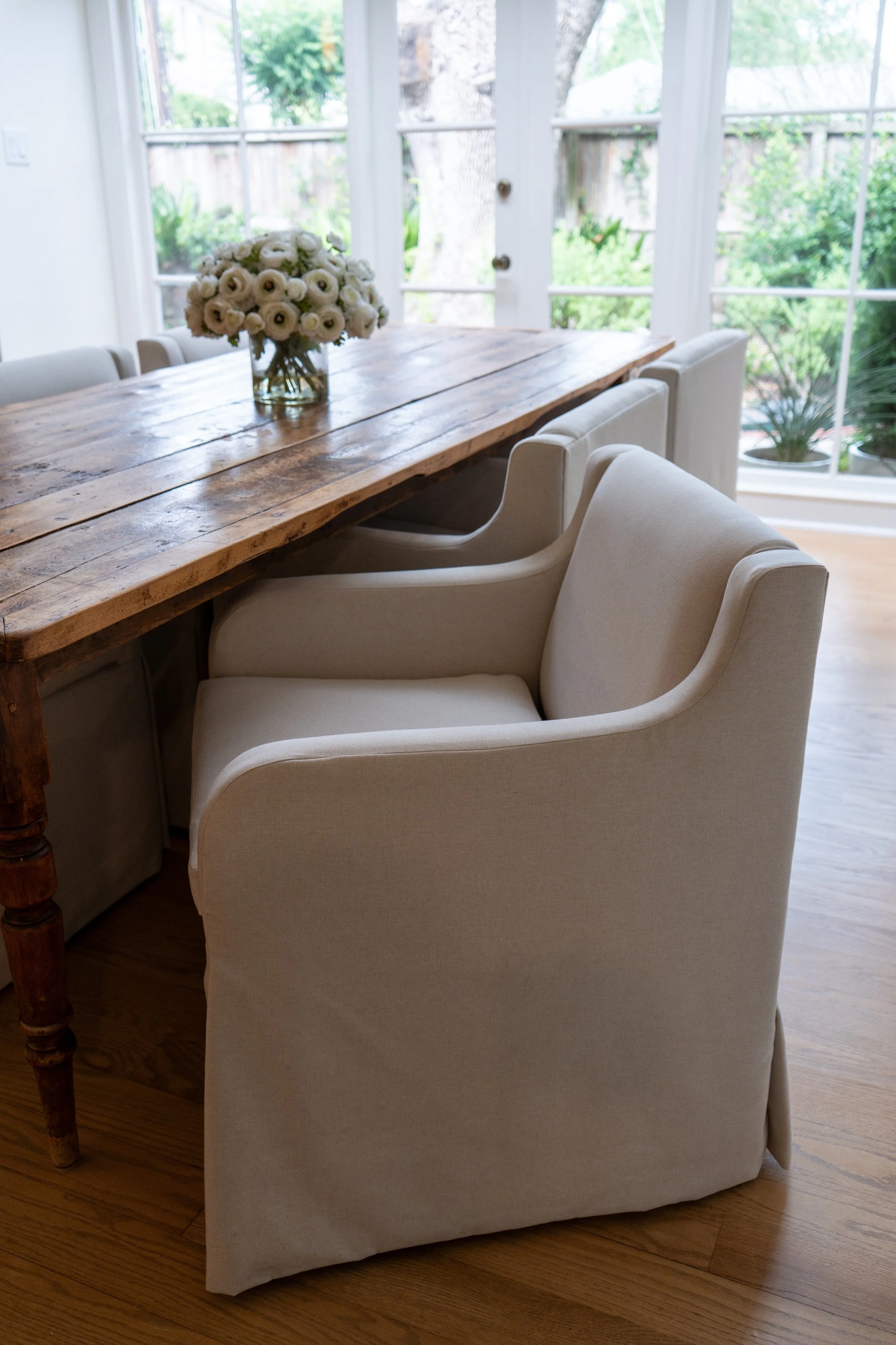 A dining room with a rustic wooden table, white upholstered chairs, a large glass vase with white flowers, and large windows showing greenery outside.