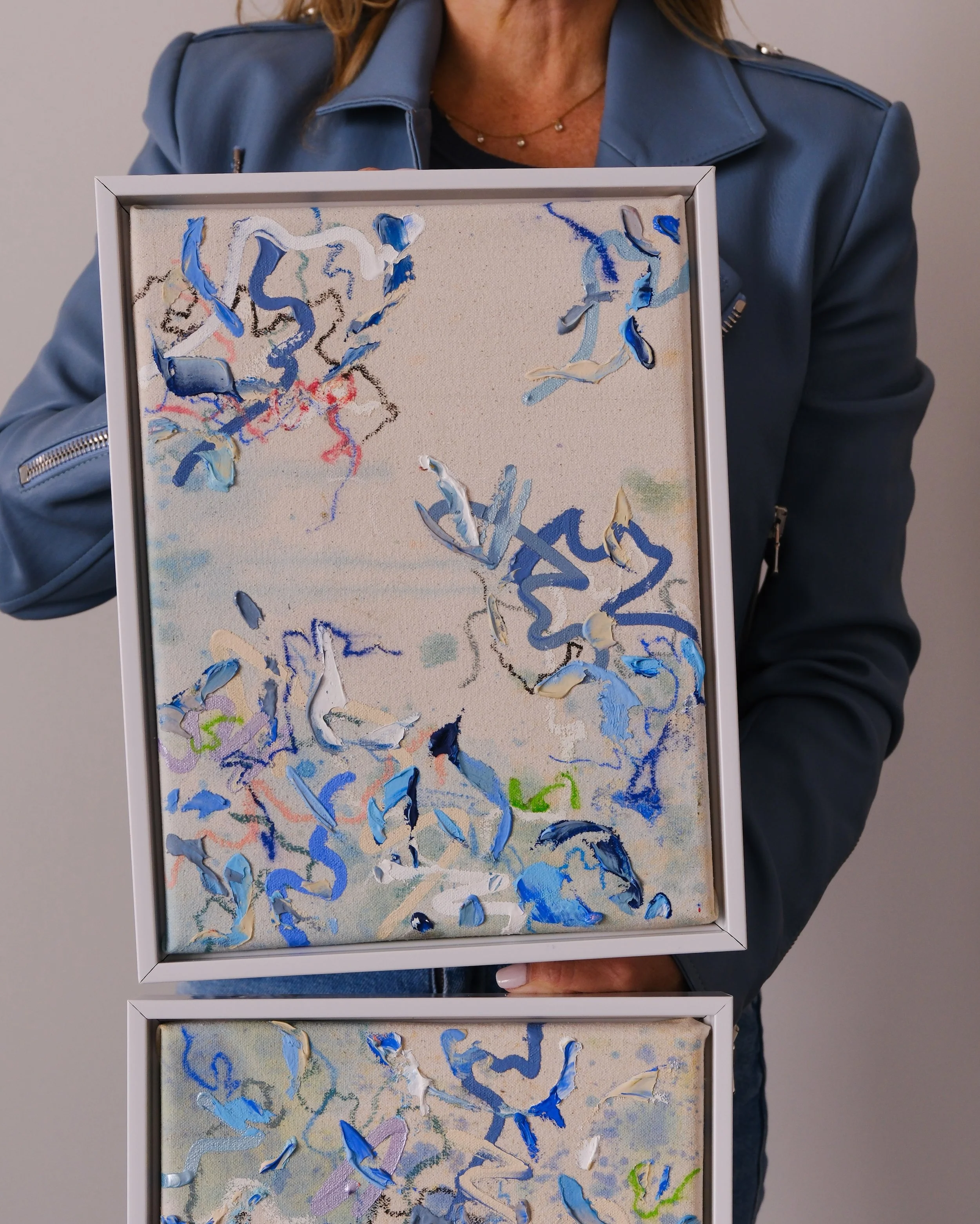 A person wearing a blue jacket holding two framed abstract paintings with thick, swirling strokes of various shades of blue, white, and other pastel colors.