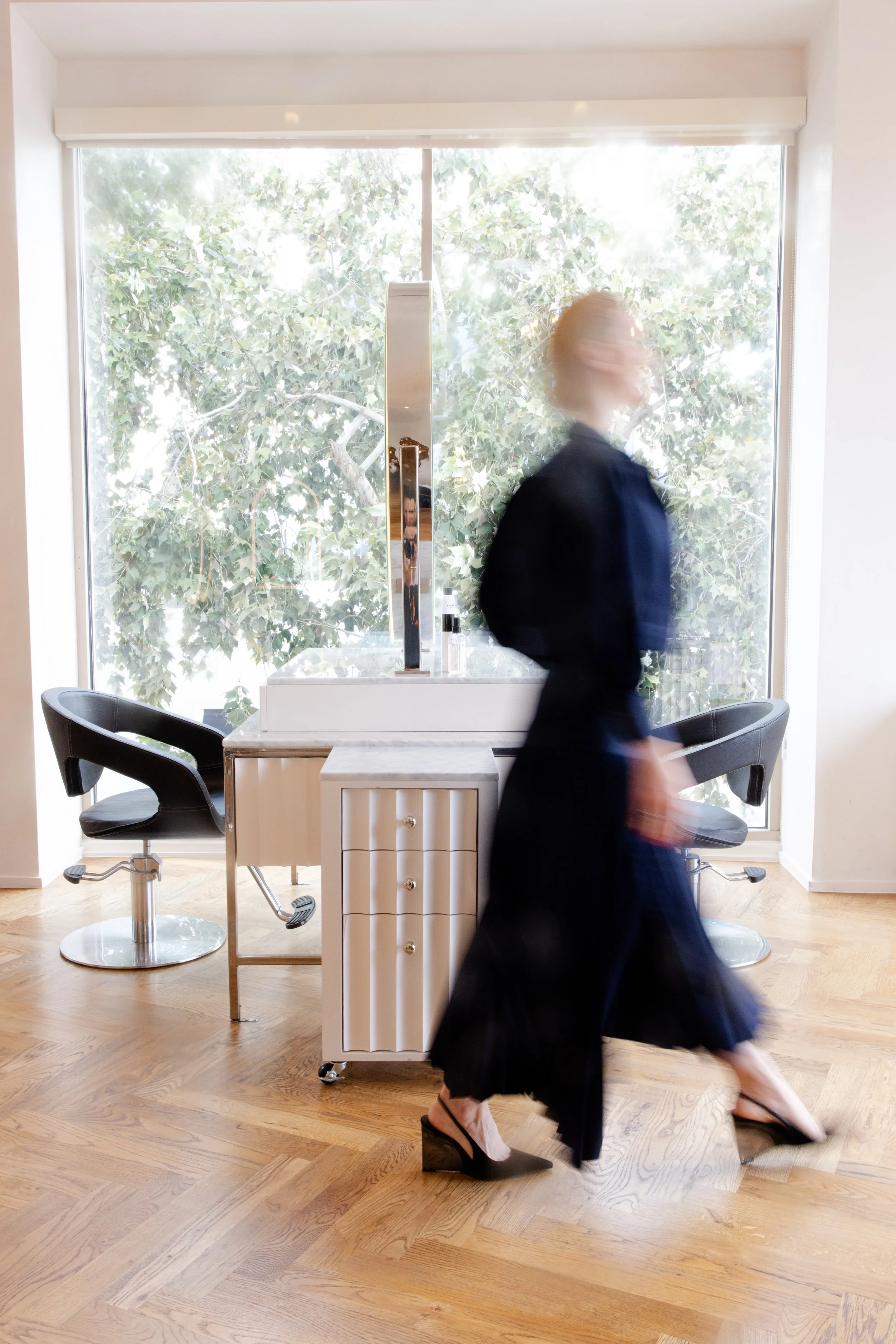 A person in a black dress and white high heels walking past a salon chair and a small cabinet, with a large window showing trees outside in the background.
