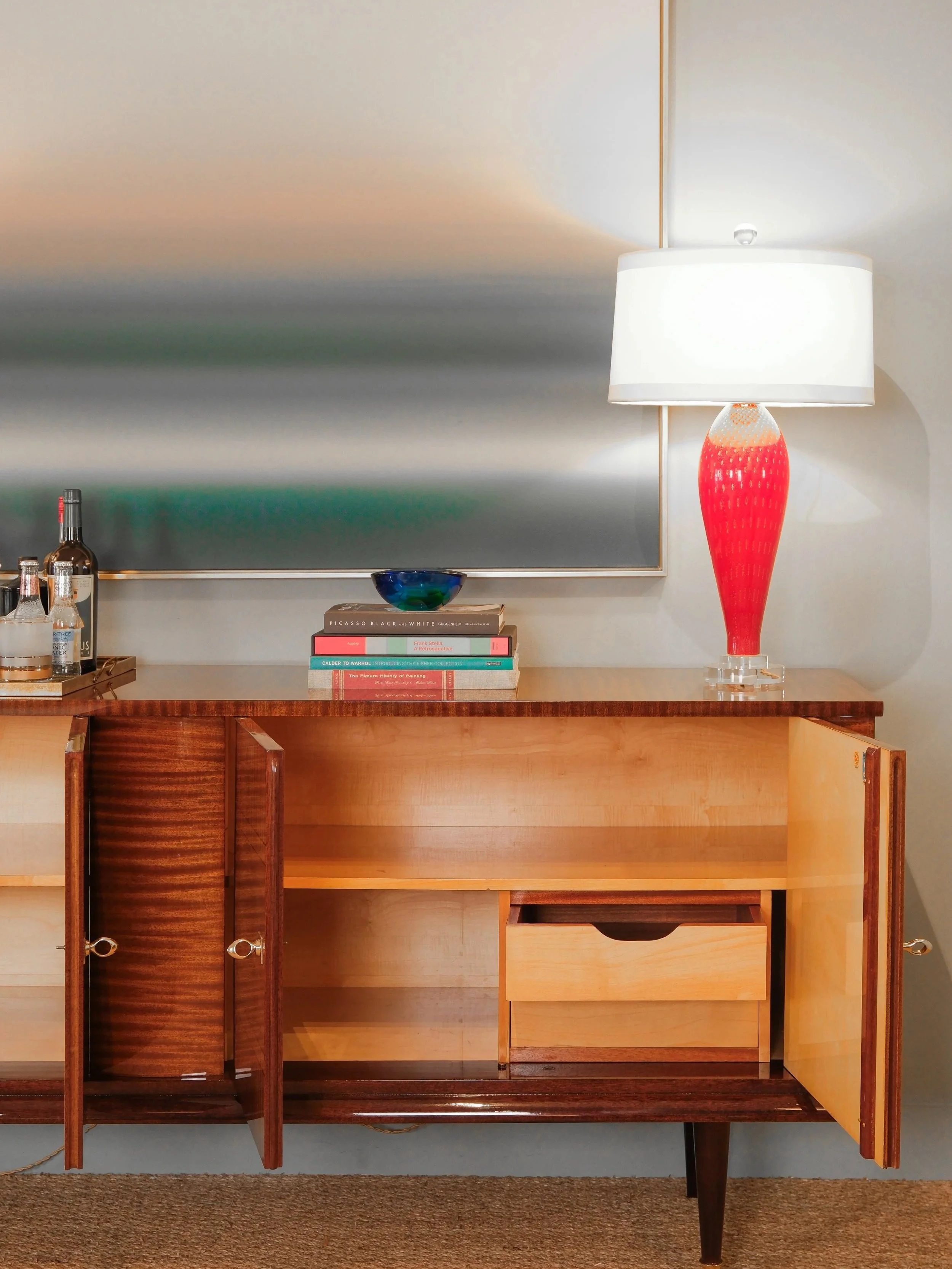 A mid-century modern wooden sideboard with a white and red glass lamp, several bottles, and a stack of art books on top, positioned against a wall with a large landscape painting.