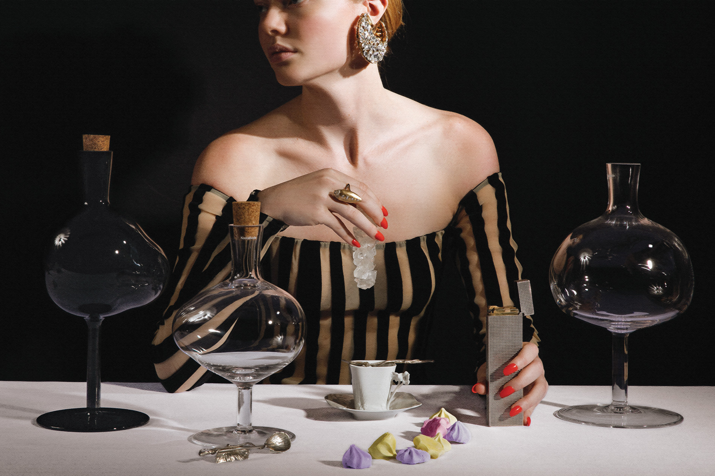A woman with red hair wearing a black-and-gold striped off-the-shoulder top and large earrings sits at a table with dark bottles, glasses, a cup with a dessert, and scattered colorful meringues against a black background.
