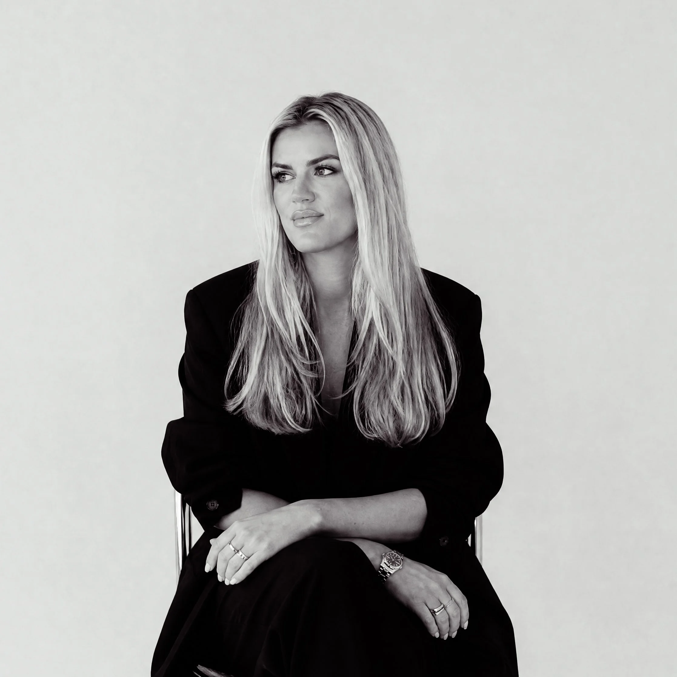 Black-and-white portrait of a woman with long blonde hair, wearing a black blazer, sitting with her hand resting on her knee, against a plain light background.